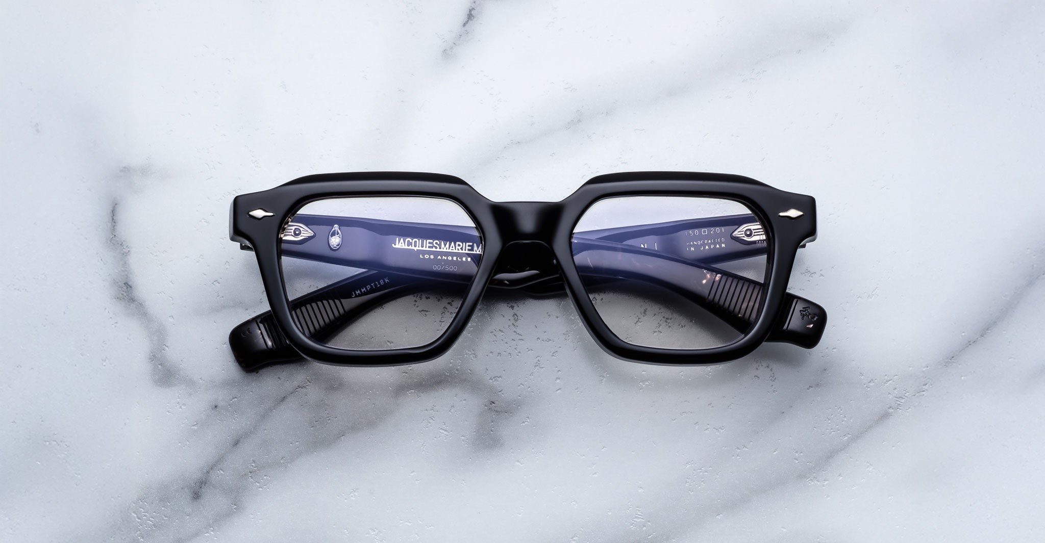 A pair of Jacques Marie Mage Tomlin black rectangular eyeglasses with thick frames rests on a white marble surface. The lenses show a subtle blue reflection, and the brand name appears on the inner left arm.
