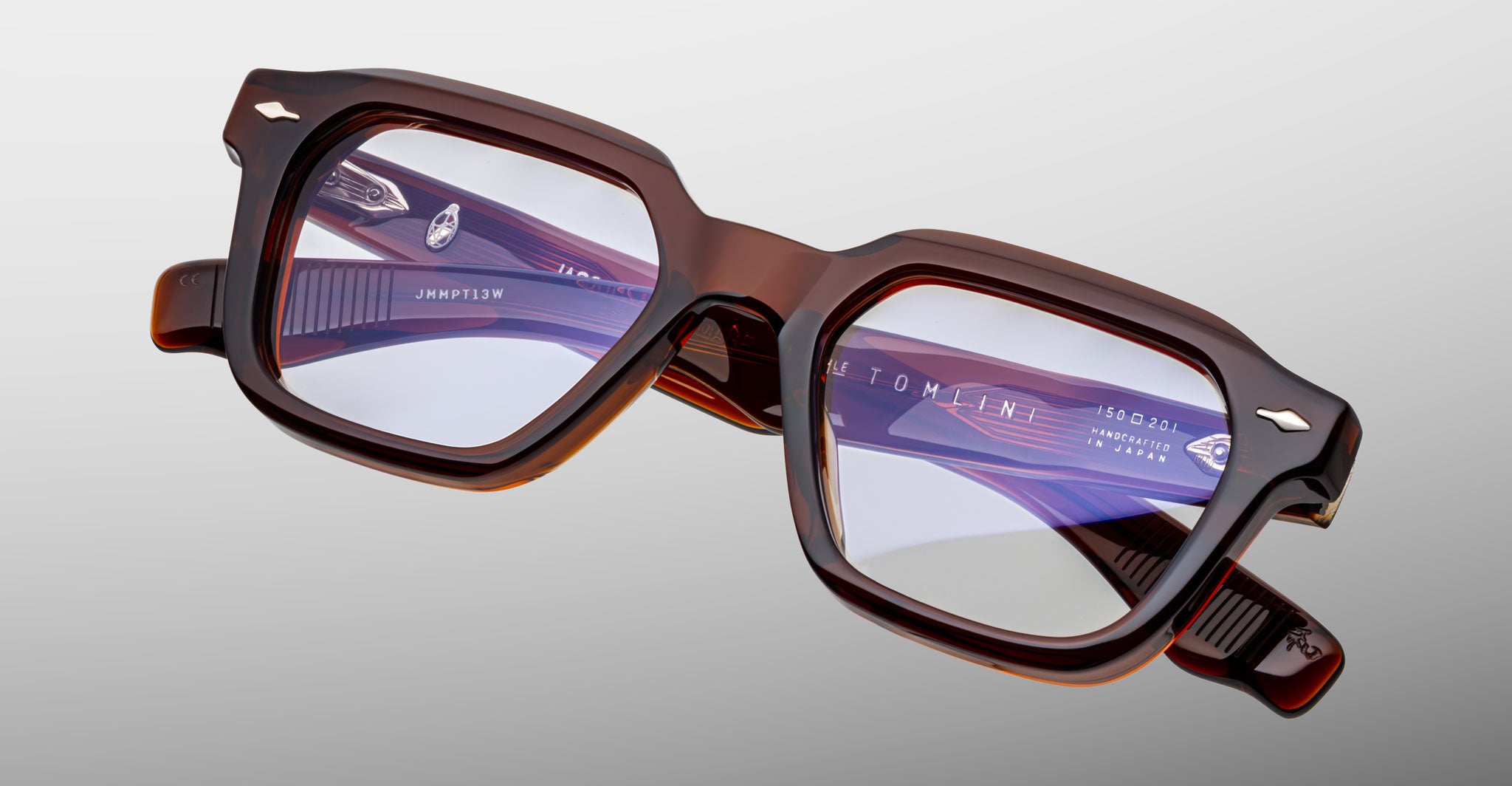 A pair of Jacques Marie Mage Tomlin brown, thick-rimmed rectangular eyeglasses with clear lenses rests on a light gray surface. White text with branding and model details appears inside one arm.