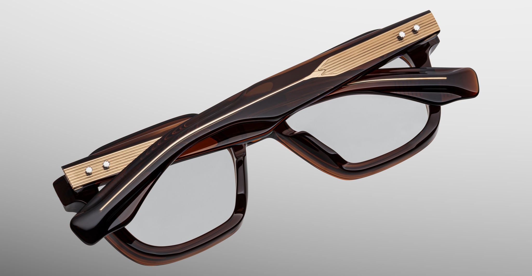The Jacques Marie Mage Tomlin rectangular eyeglasses feature a dark brown frame with gold metal temple accents, shown closed and viewed from above on a light gray background.