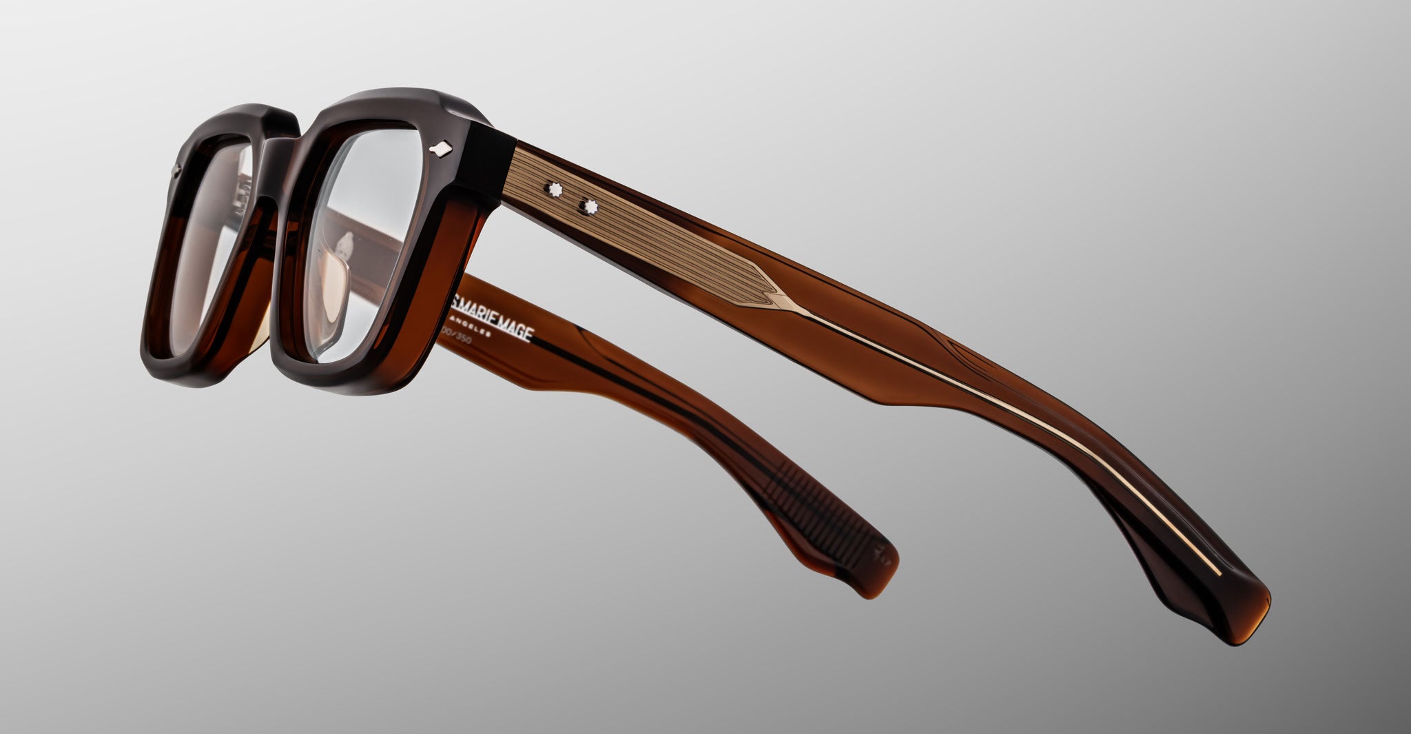 The Jacques Marie Mage Tomlin eyeglasses feature brown rectangular thick frames, gold temple detailing, two small metal rivets on each side, and clear lenses, displayed against a gradient gray background.