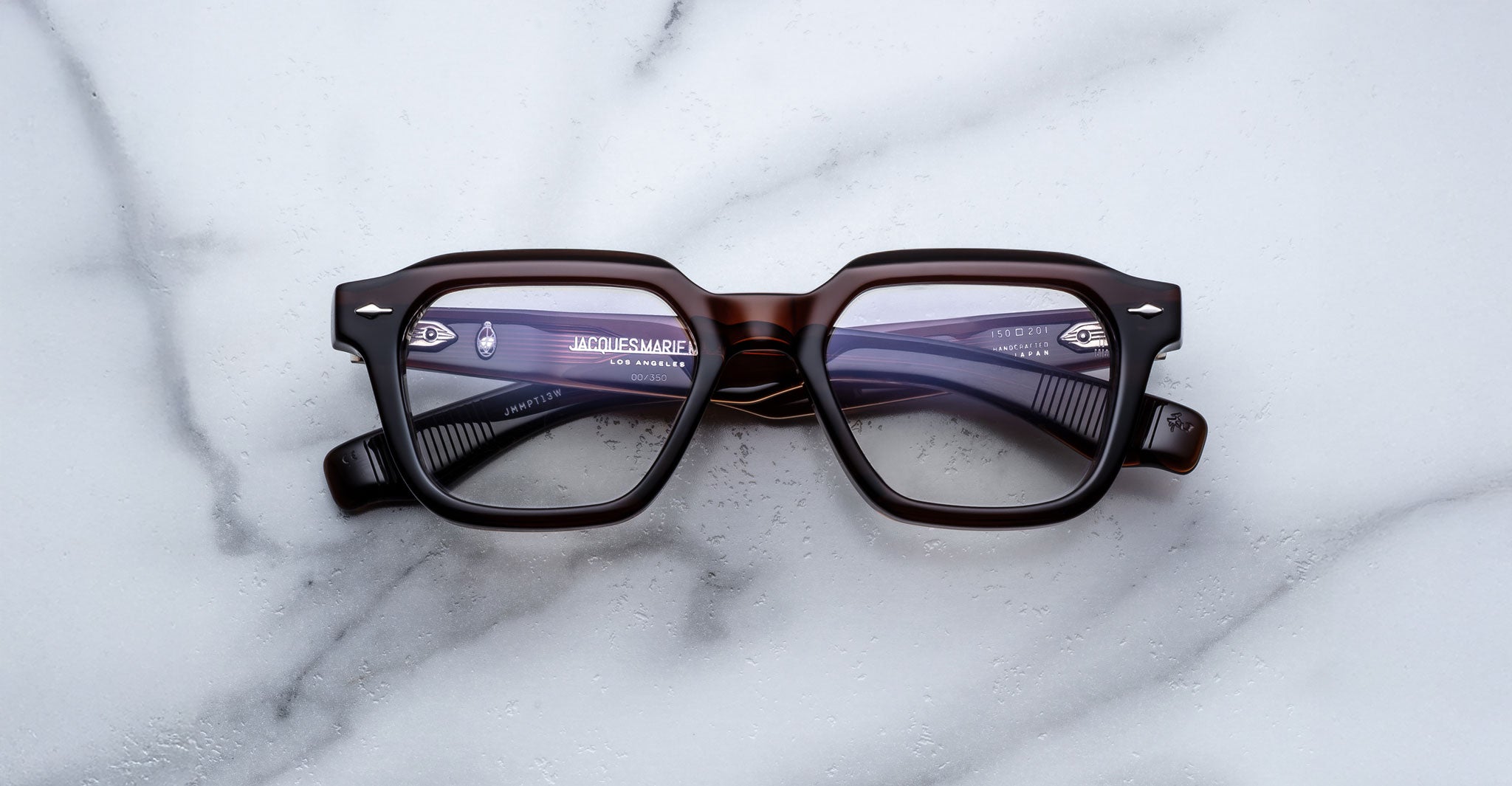 Jacques Marie Mage Tomlin thick, dark-brown rectangular eyeglass frames with clear lenses rest on a white marble surface. Branding text and logos are visible on the inside arms of the glasses.