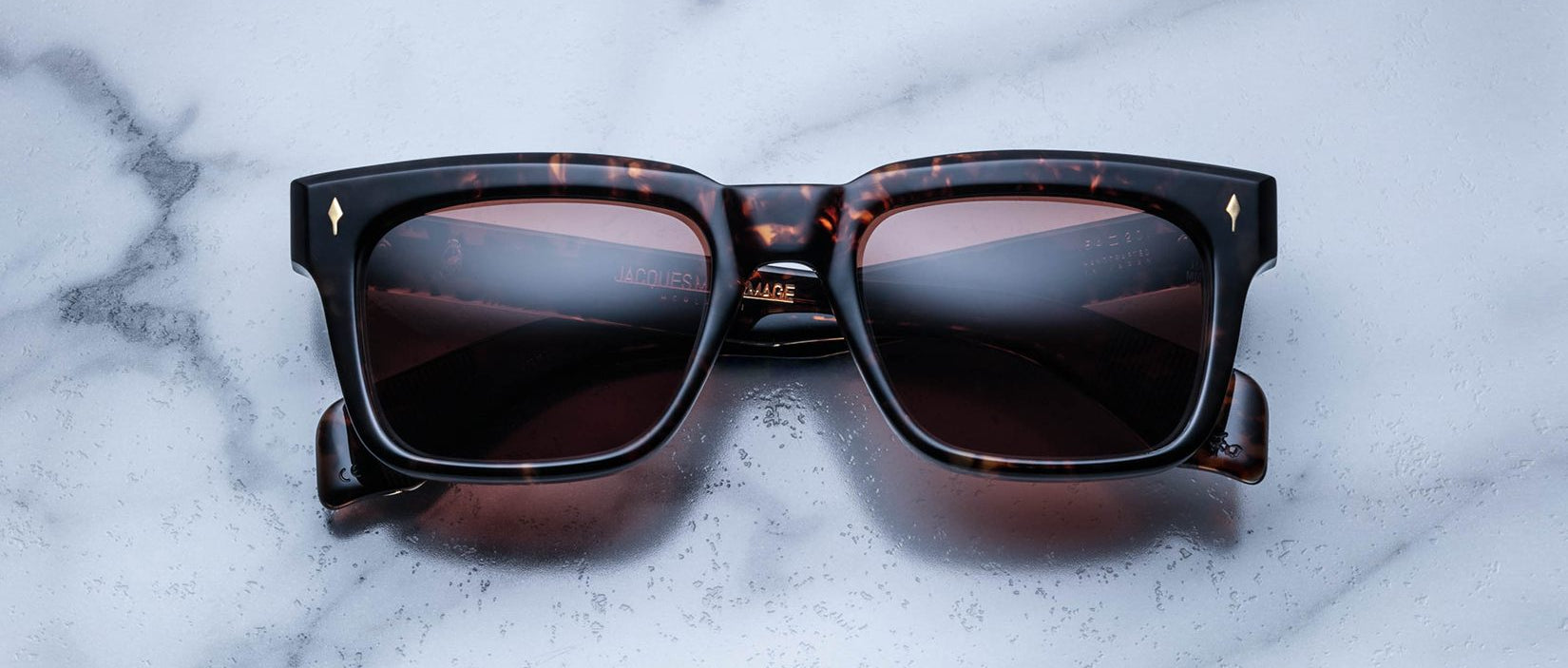 The Jacques Marie Mage Torino eyeglasses, featuring bold rectangular dark tortoiseshell acetate frames and dark lenses, handcrafted by Jacques Marie Mage, rest on a white marble surface.