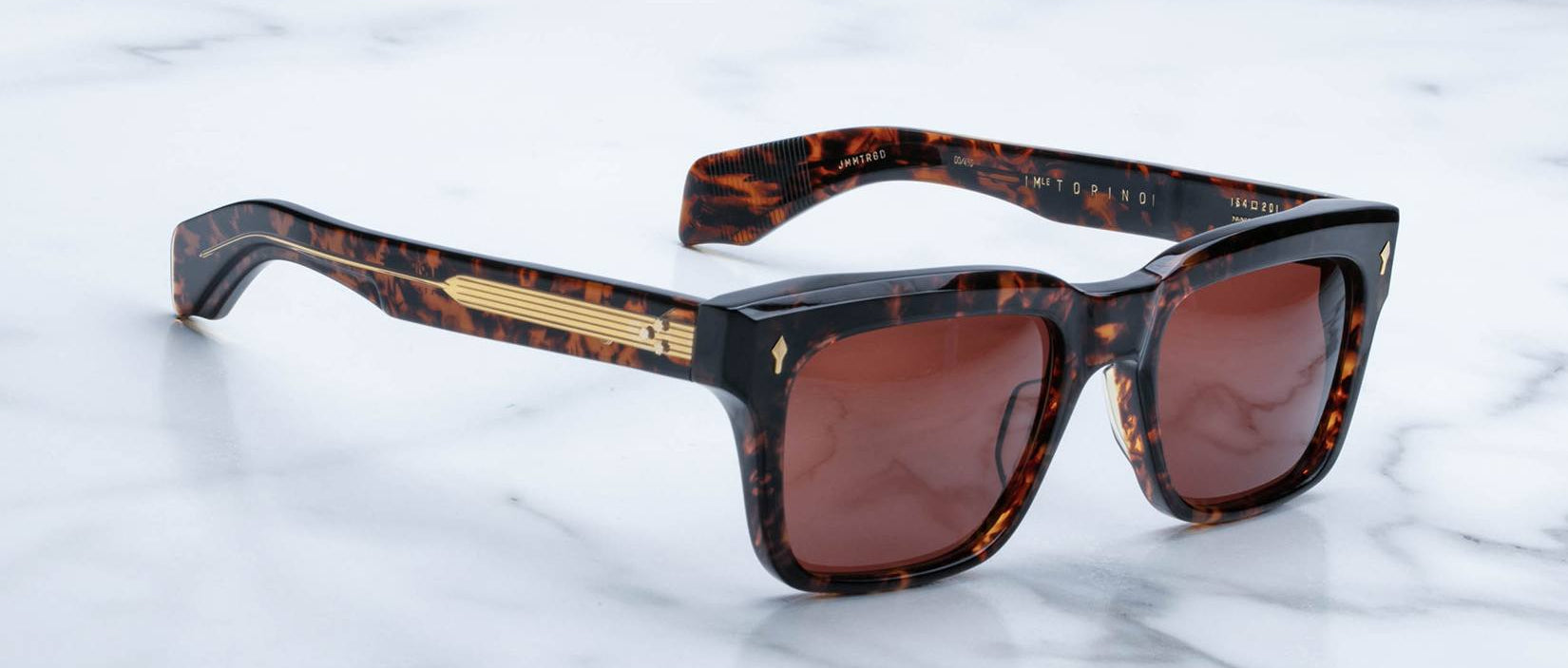 Jacques Marie Mage Torino sunglasses by Jacques Marie Mage feature bold tortoiseshell rectangular acetate frames, brown lenses, and gold accents on the arms, shown resting on a white marble surface.