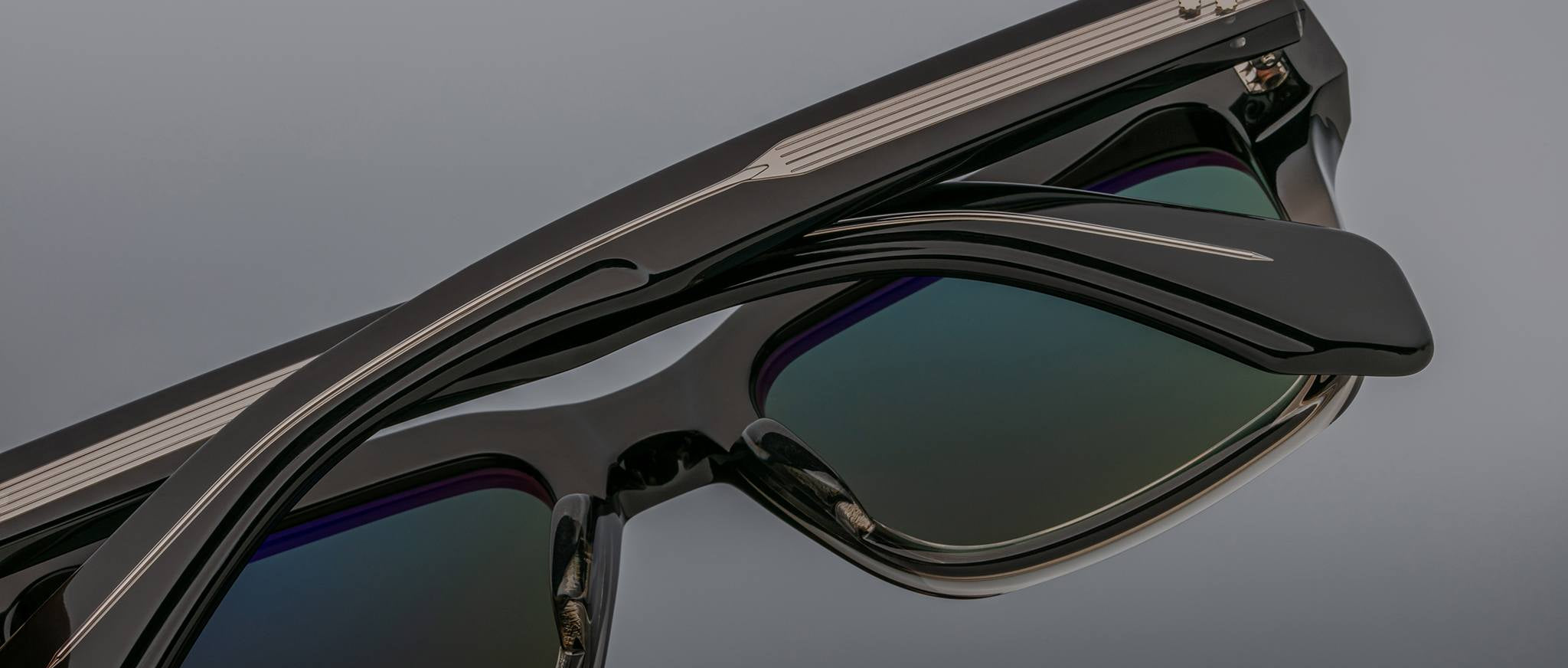 Close-up of Jacques Marie Mage Torino eyeglass frames in bold black with dark reflective lenses and silver arm details, set against a gray background.