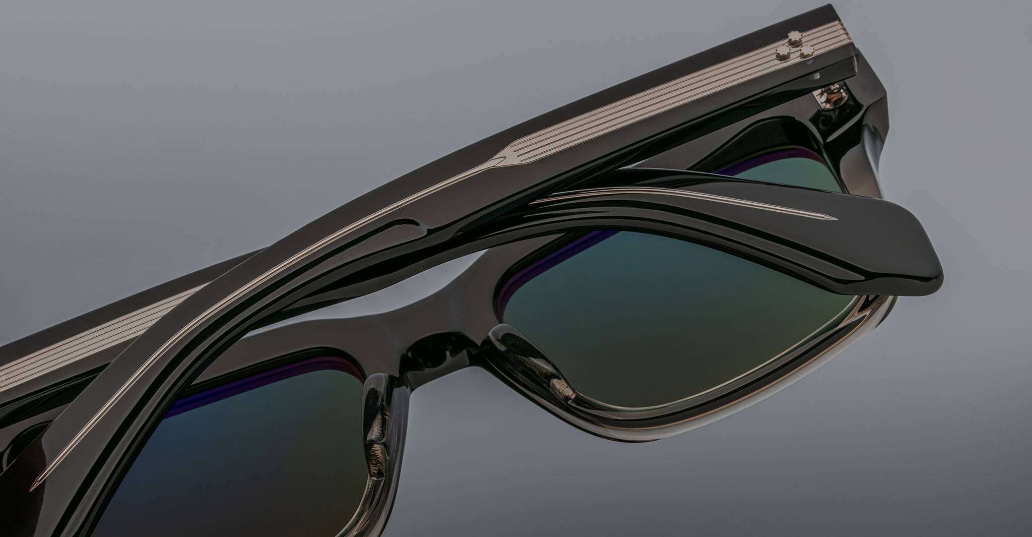Close-up of Jacques Marie Mage Torino eyeglass frames in bold black with dark reflective lenses and silver arm details, set against a gray background.