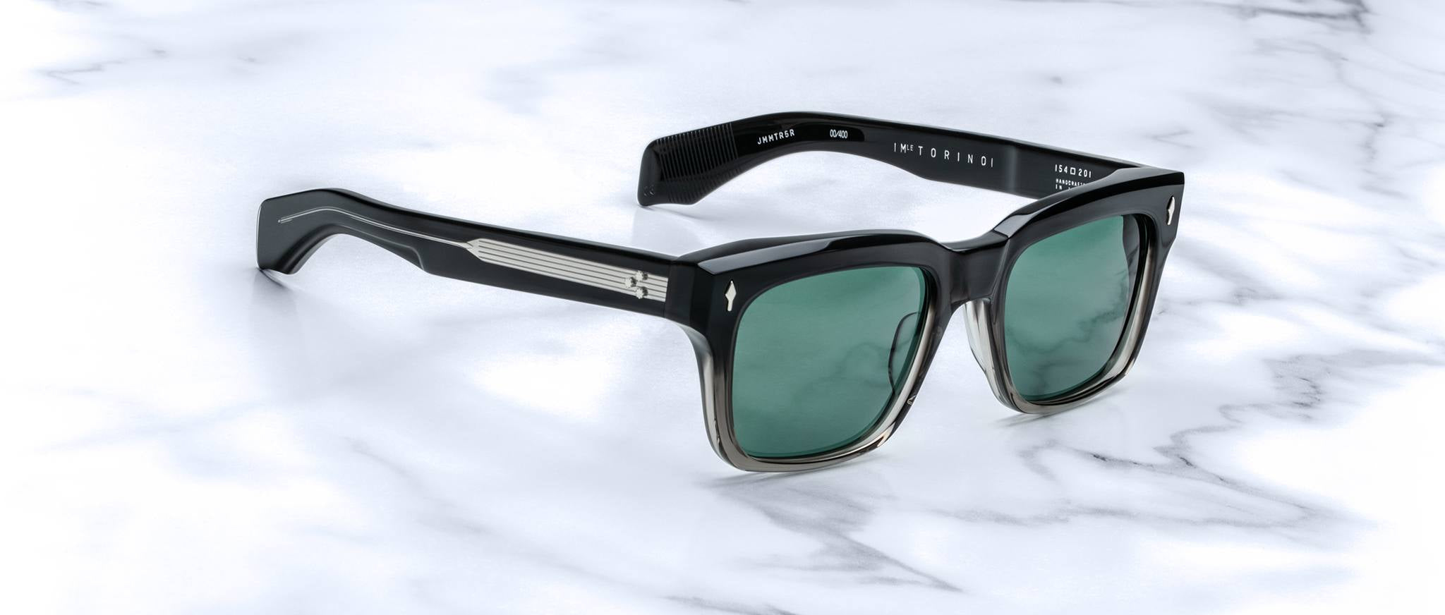 Jacques Marie Mage Torino by Jacques Marie Mage features black bold rectangular acetate frames, green lenses, and silver arm detailing, beautifully displayed on a white marble surface.
