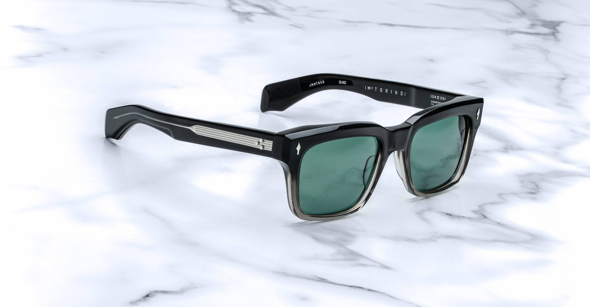 Jacques Marie Mage Torino by Jacques Marie Mage features black bold rectangular acetate frames, green lenses, and silver arm detailing, beautifully displayed on a white marble surface.