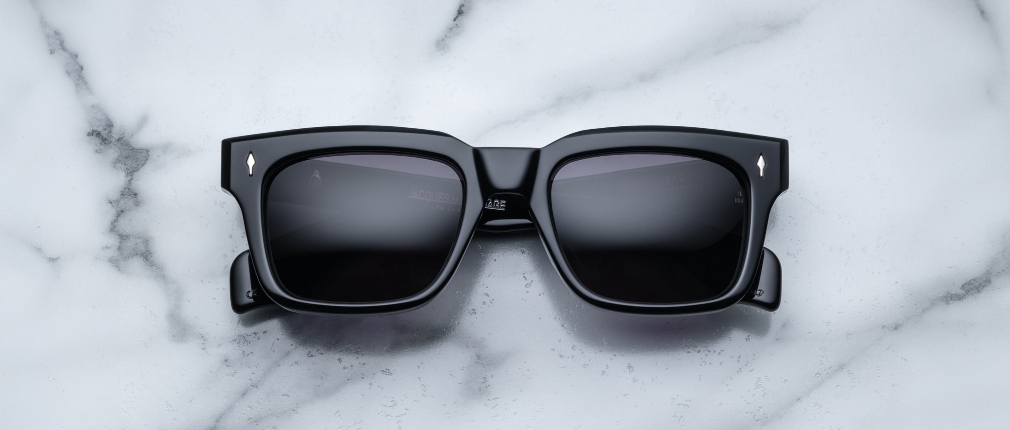 The Jacques Marie Mage Torino rectangular sunglasses feature bold handcrafted acetate frames and dark lenses, resting on a white marble surface for a classic look.