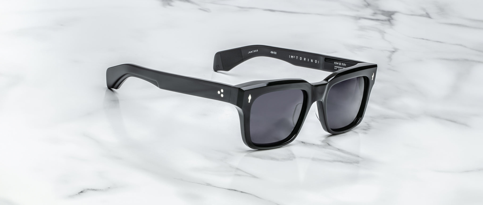 The Jacques Marie Mage Torino sunglasses by Jacques Marie Mage feature bold black rectangular frames and dark lenses, elegantly displayed on a white marble surface.