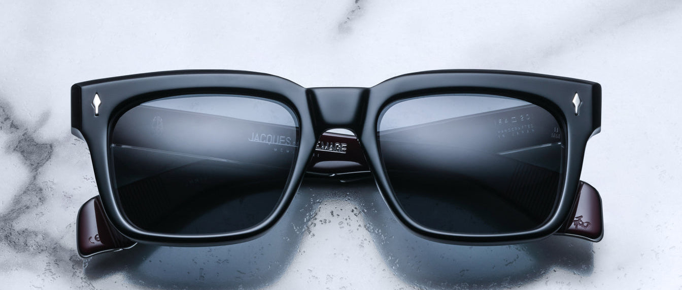 The Jacques Marie Mage Torino sunglasses by Jacques Marie Mage feature bold black rectangular frames and dark lenses, displayed on a white marble surface.