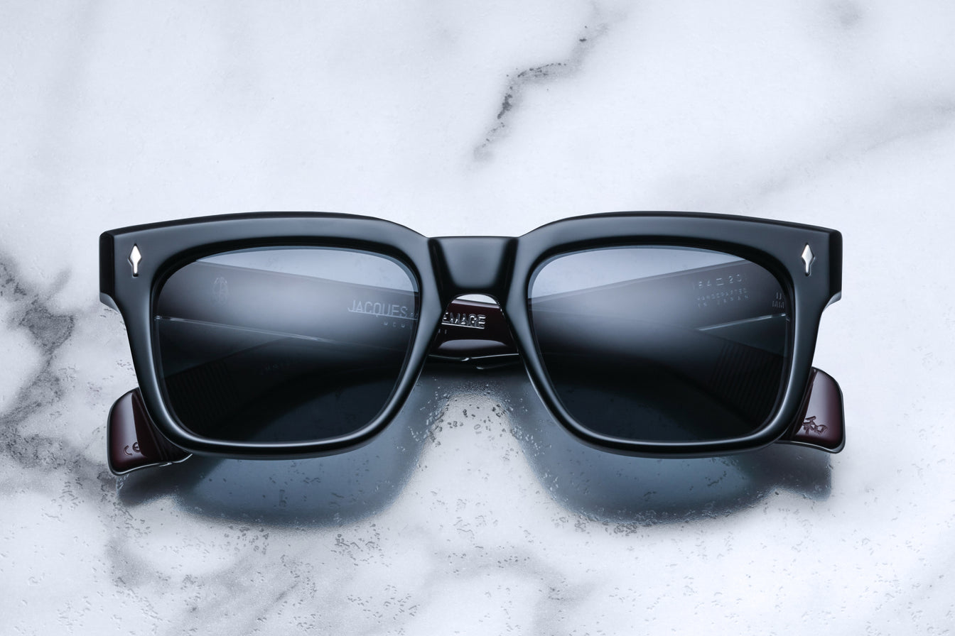 The Jacques Marie Mage Torino sunglasses by Jacques Marie Mage feature bold black rectangular frames and dark lenses, displayed on a white marble surface.