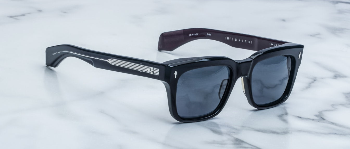 Jacques Marie Mage Torino bold rectangular acetate eyeglass frames with dark lenses and metallic accents rest on a white marble surface.
