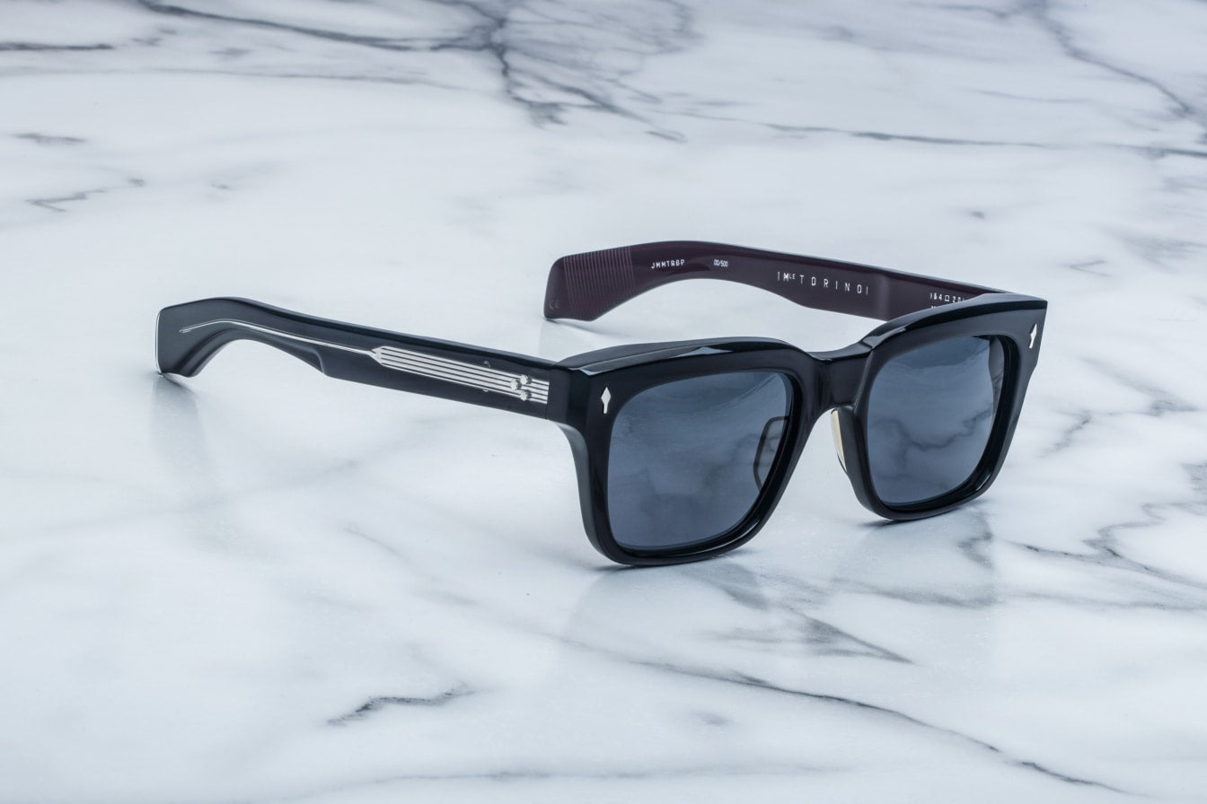Jacques Marie Mage Torino bold rectangular acetate eyeglass frames with dark lenses and metallic accents rest on a white marble surface.