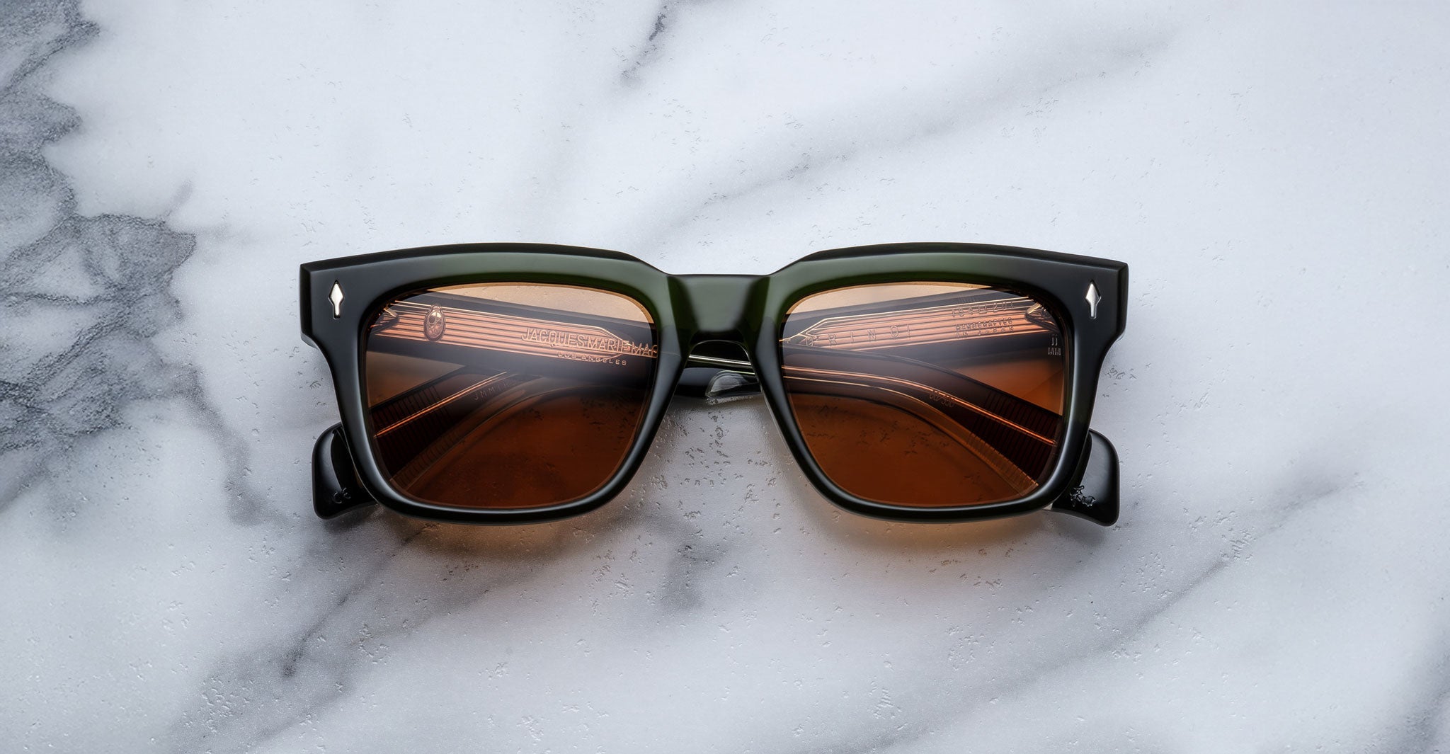 The Jacques Marie Mage Torino sunglasses feature bold, square black acetate frames and brown tinted lenses, displayed on a white marble surface that reflects patterns of light, showcasing the brand's timeless handcrafted style.