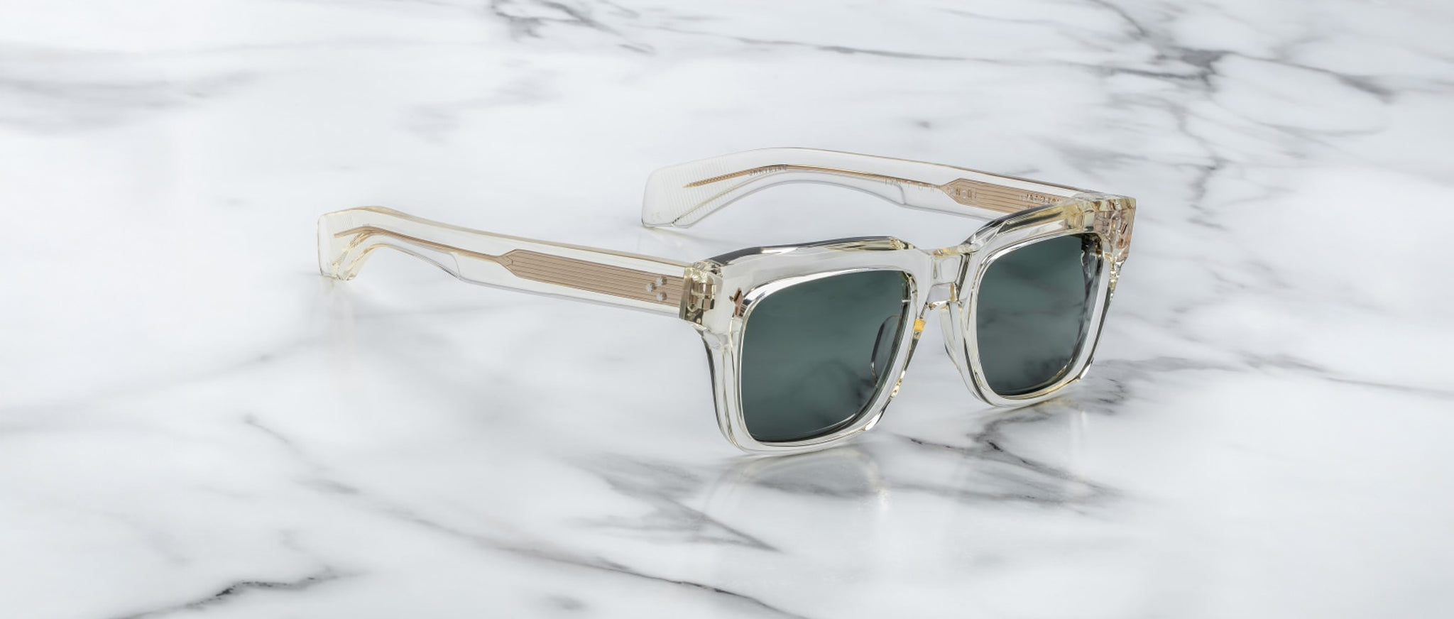 Clear-framed Jacques Marie Mage Torino sunglasses with dark lenses, inspired by bold rectangular designs, rest on white marble with gray veining.
