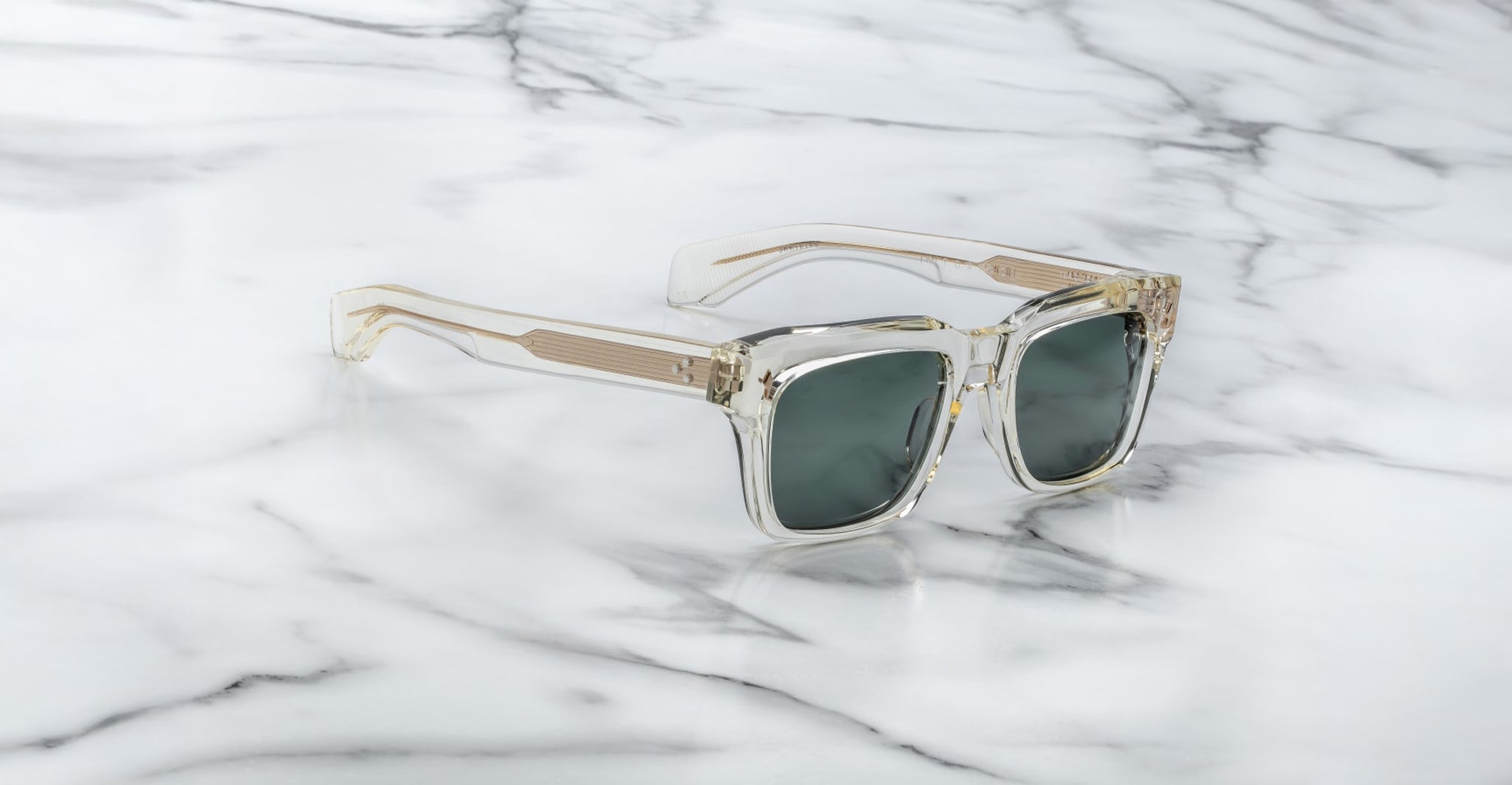 Clear-framed Jacques Marie Mage Torino sunglasses with dark lenses, inspired by bold rectangular designs, rest on white marble with gray veining.