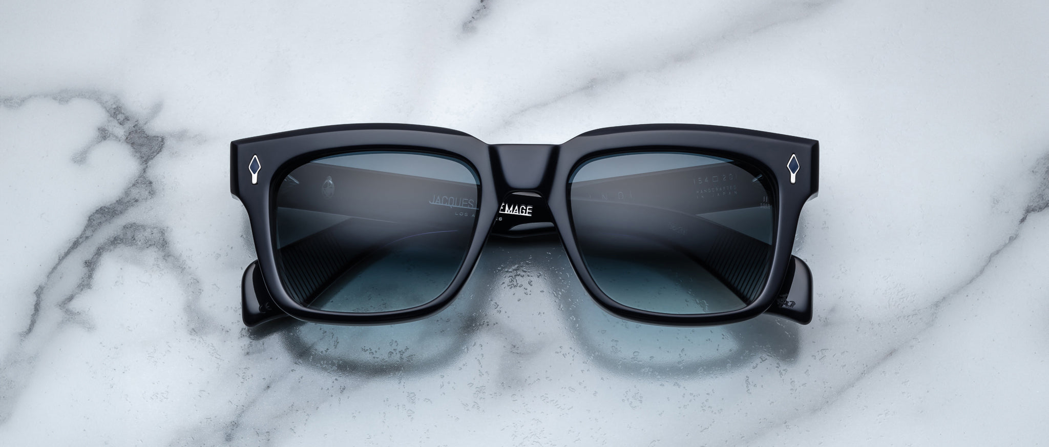 The Jacques Marie Mage Torino by Jacques Marie Mage are black rectangular sunglasses with dark tinted lenses and bold frames, displayed on a white marble surface.