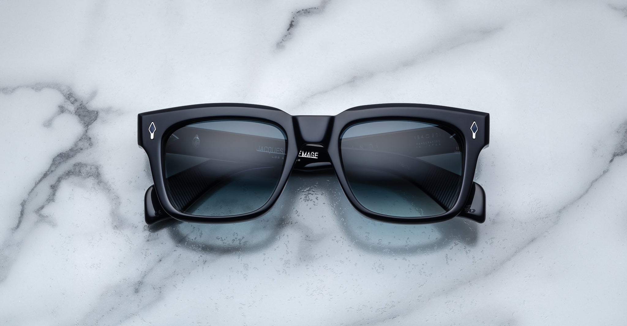The Jacques Marie Mage Torino by Jacques Marie Mage are black rectangular sunglasses with dark tinted lenses and bold frames, displayed on a white marble surface.