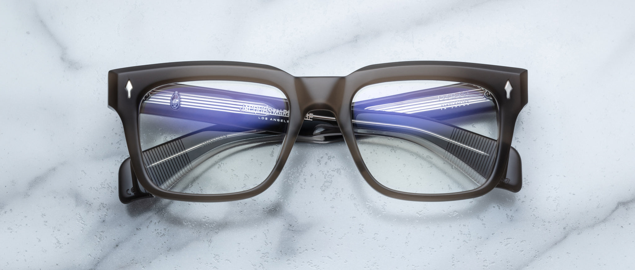 A pair of bold, rectangular Jacques Marie Mage Torino eyeglass frames rests on a white marble surface.