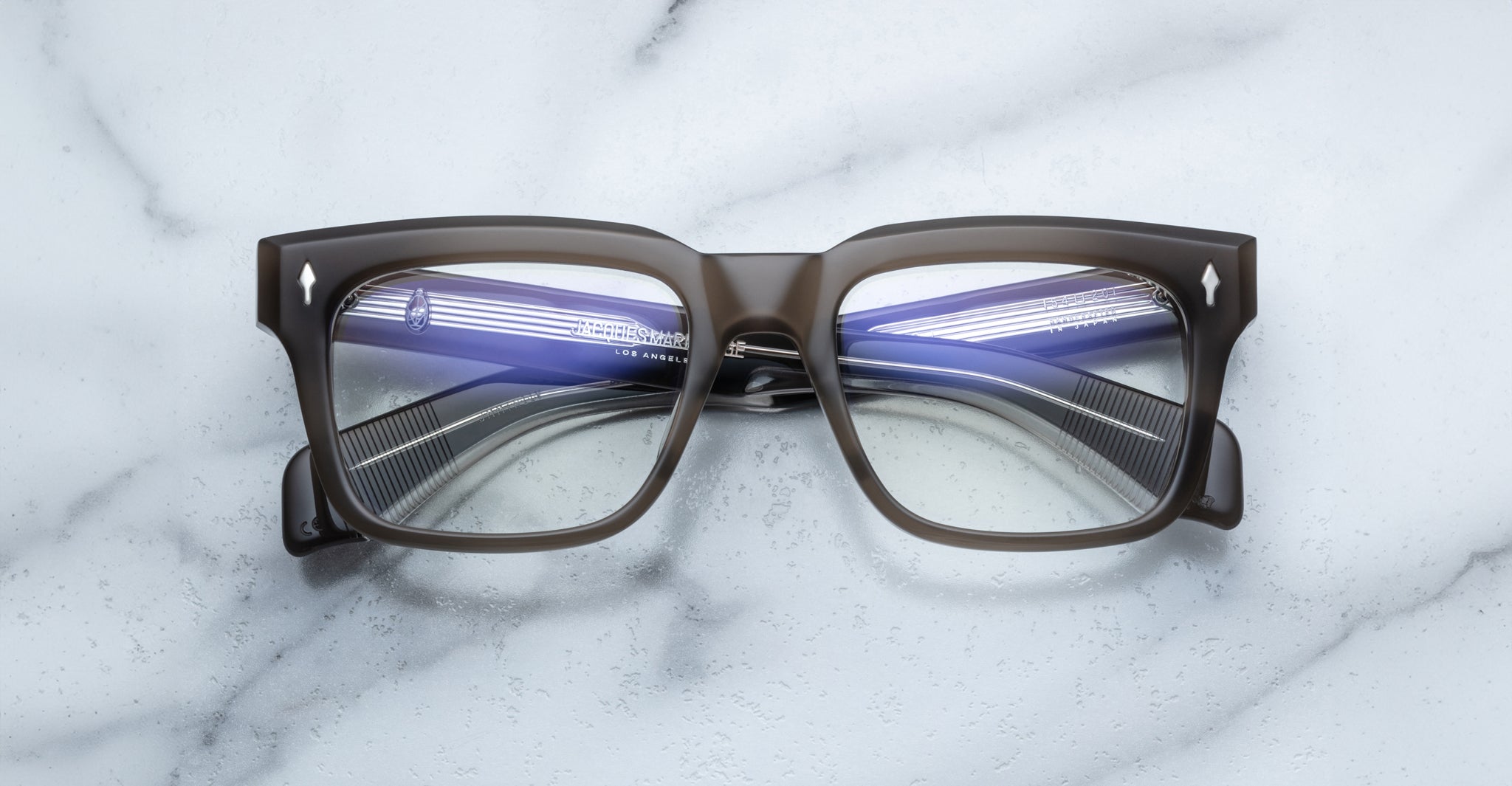 A pair of bold, rectangular Jacques Marie Mage Torino eyeglass frames rests on a white marble surface.
