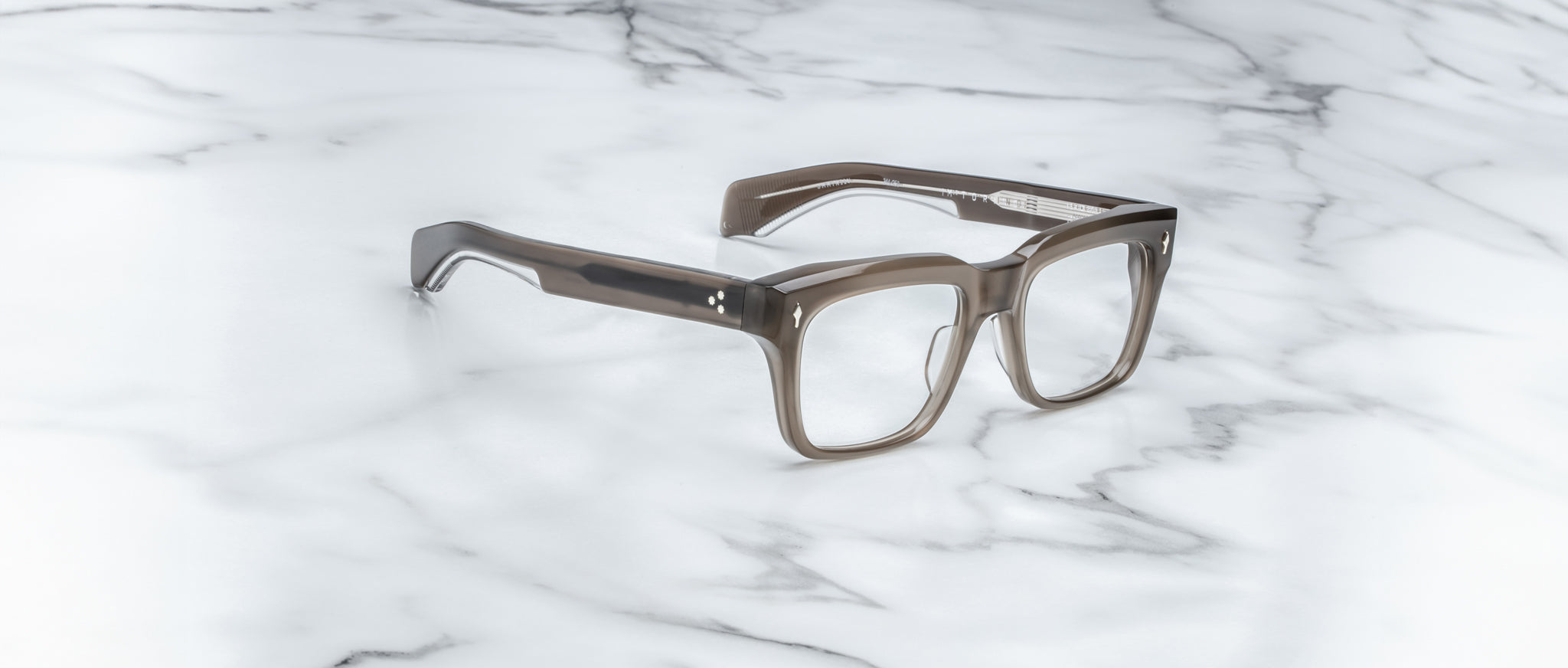 The Jacques Marie Mage Torino handcrafted acetate eyewear features bold gray translucent rectangular frames, elegantly displayed on a white marble surface.