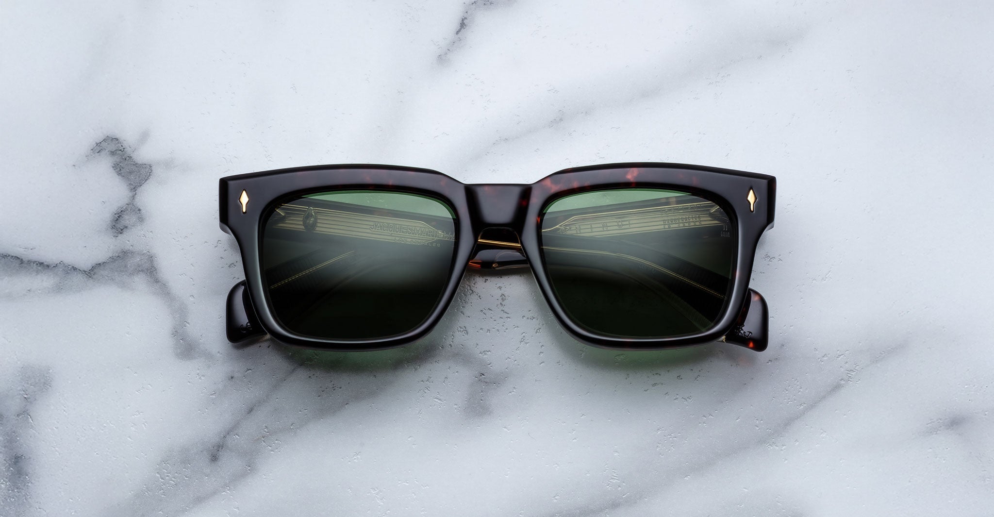A pair of Jacques Marie Mage Torino rectangular eyeglass frames, handcrafted from Japanese acetate with green lenses, rests on a white marble surface.