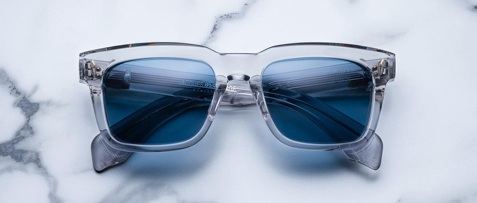 Jacques Marie Mage Torino sunglasses feature clear frames and blue-tinted lenses, inspired by the brand's bold rectangular design, displayed on a white marble surface.