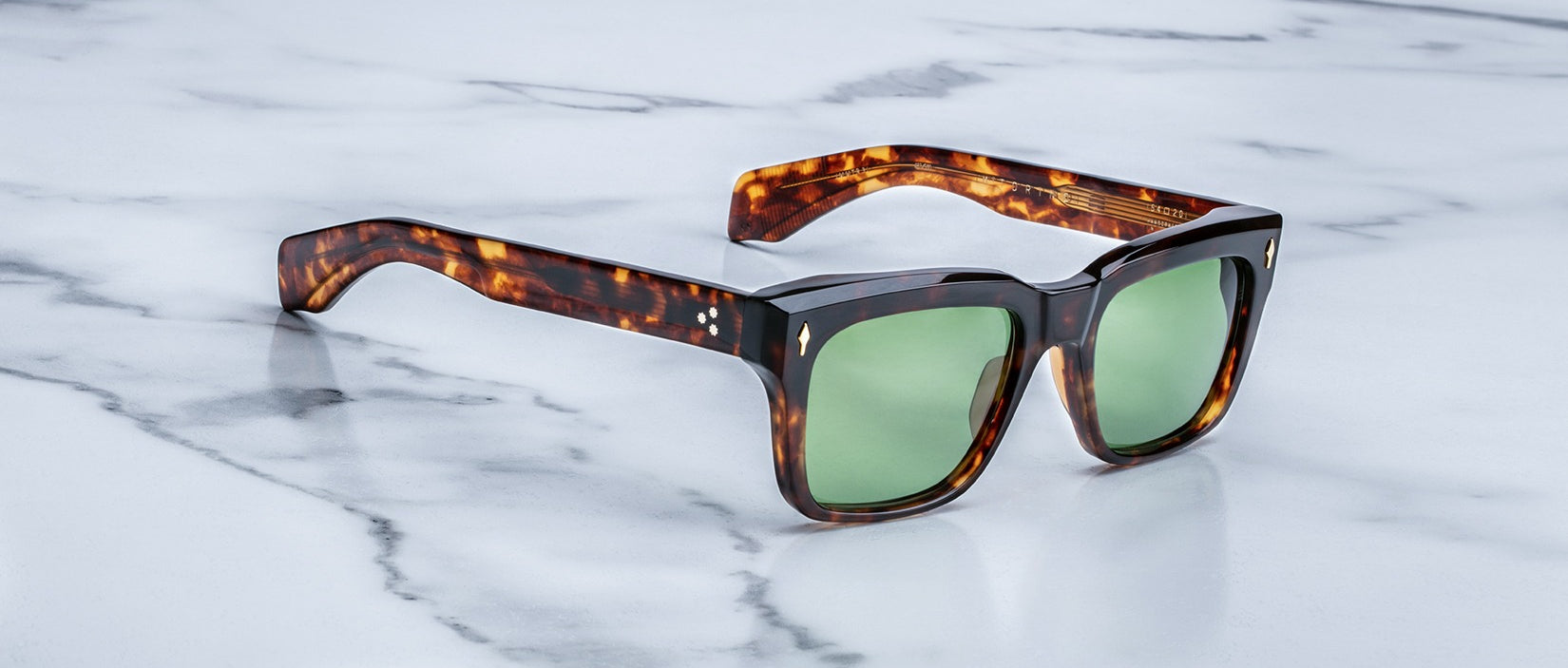 The Jacques Marie Mage Torino handcrafted acetate sunglasses in tortoiseshell with green lenses are displayed on a white marble surface.