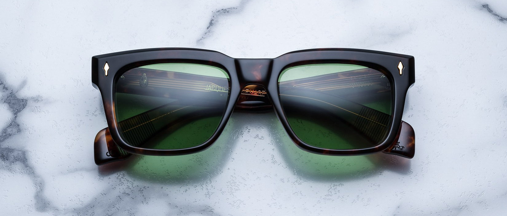 The Jacques Marie Mage Torino eyeglass frames feature bold rectangular tortoiseshell acetate and green lenses, handcrafted by Jacques Marie Mage, shown on a white marble surface.