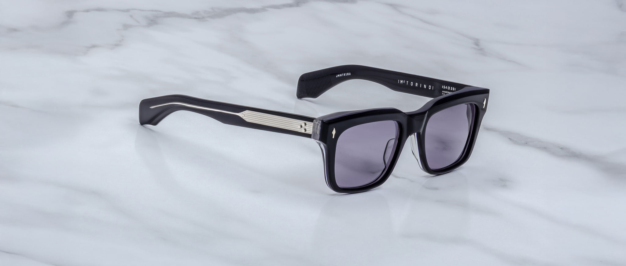 The Jacques Marie Mage Torino by Jacques Marie Mage features bold, handcrafted black acetate rectangular frames with dark lenses and silver arm detailing, displayed on a light gray marble surface.