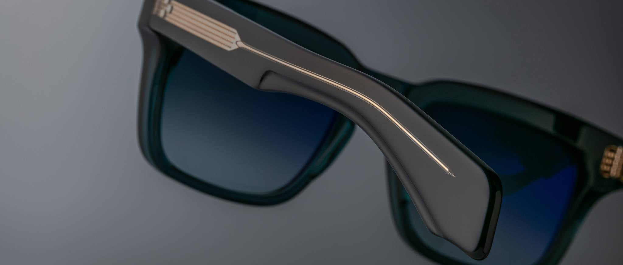 Close-up of Jacques Marie Mage Torino eyewear: bold rectangular frames with blue lenses, highlighting handcrafted acetate design details on the arm and hinge, set against a gray background.