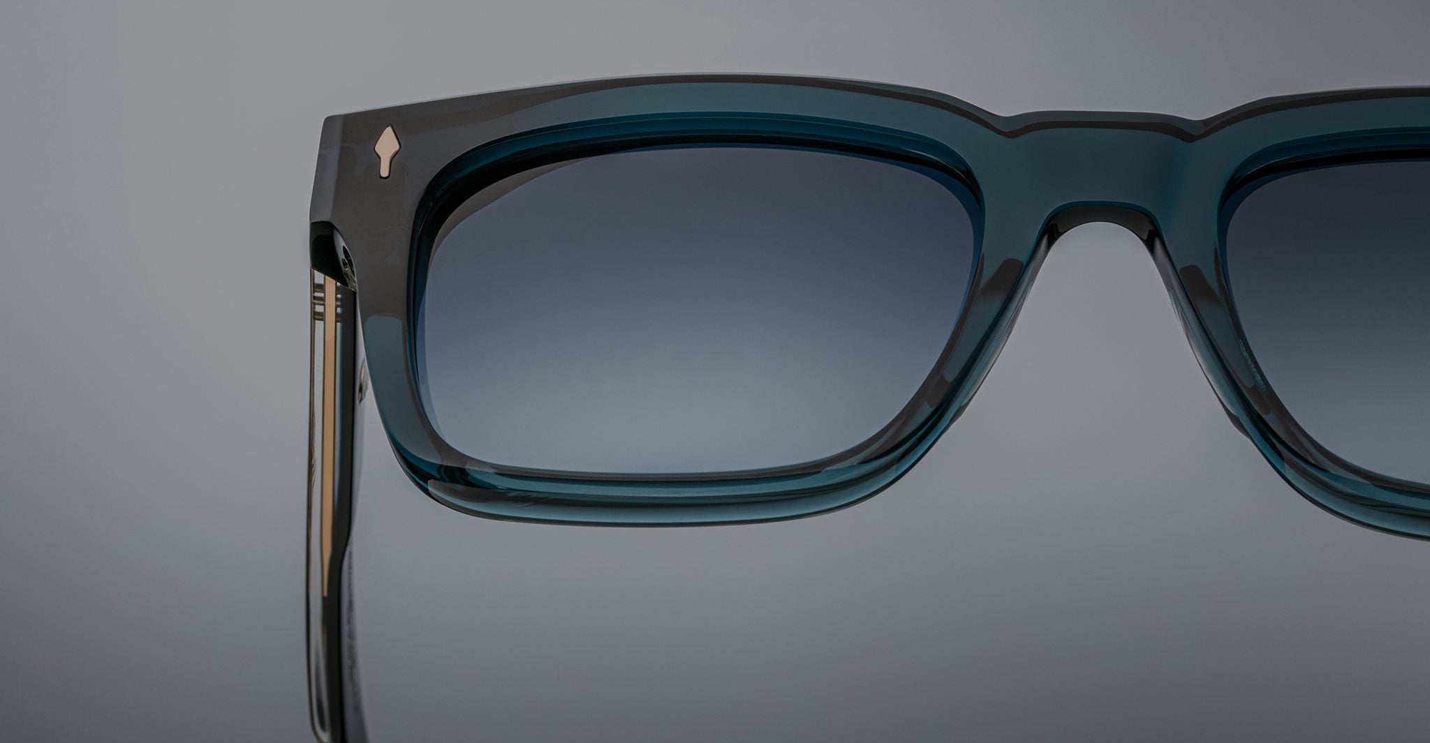 Close-up of Jacques Marie Mage Torino eyeglasses: bold, handcrafted acetate frames in teal with gradient-tinted lenses by Jacques Marie Mage, set against a plain gray background.