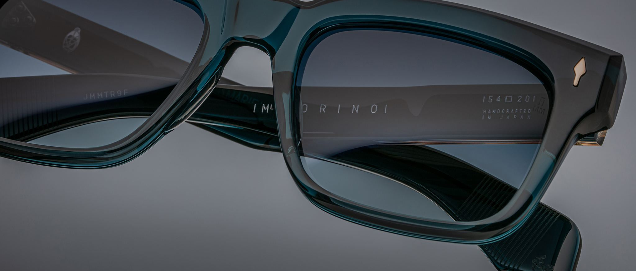 Close-up of Jacques Marie Mage Torino rectangular eyeglass frames, handcrafted acetate eyewear featuring brand markings on the inner arm, shown against a gray background.