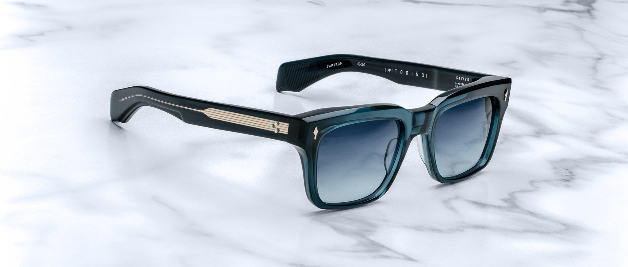 The Jacques Marie Mage Torino sunglasses feature square blue gradient lenses, bold rectangular frames, and metallic temple detailing, displayed on a white marble surface.