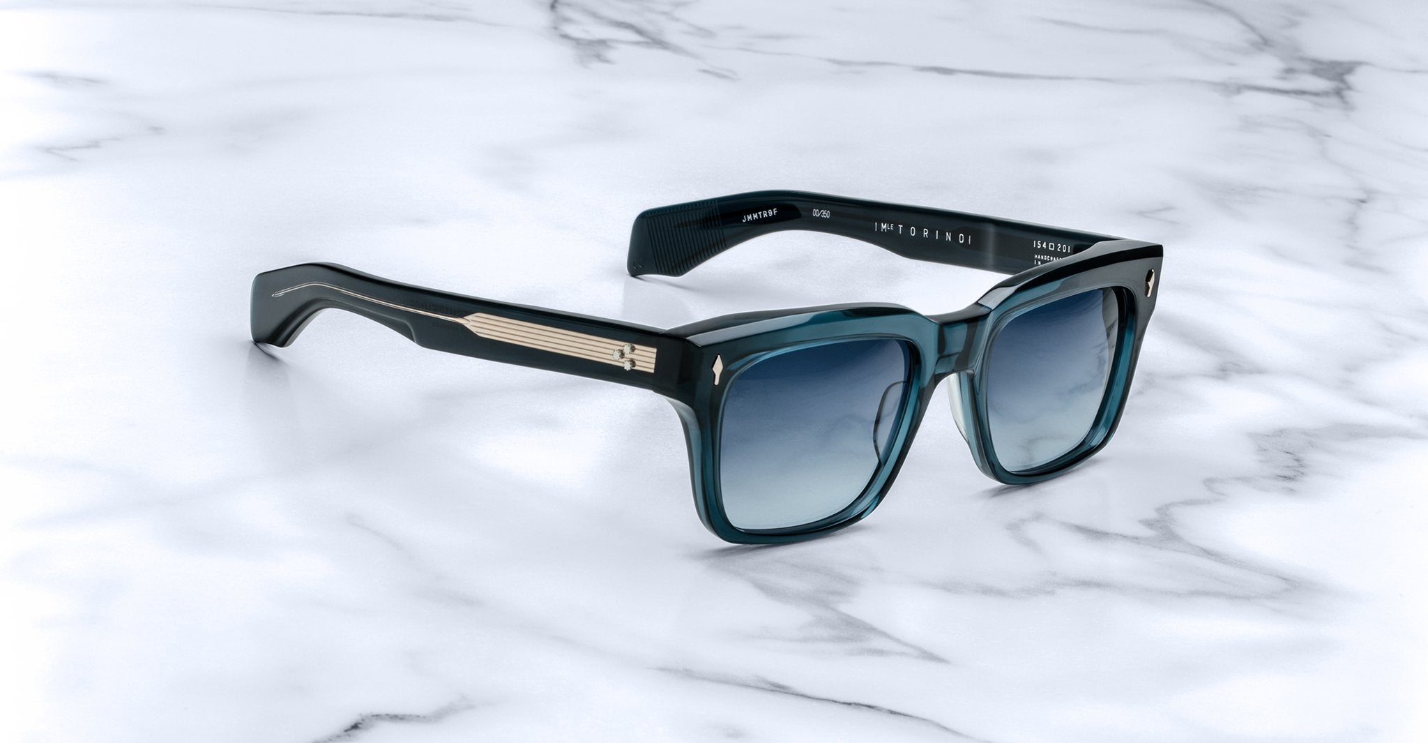 The Jacques Marie Mage Torino sunglasses feature square blue gradient lenses, bold rectangular frames, and metallic temple detailing, displayed on a white marble surface.