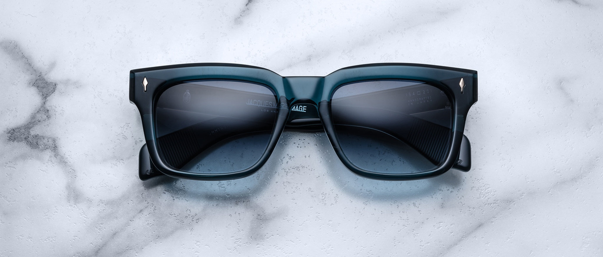 Jacques Marie Mage Torino sunglasses, featuring bold blue square acetate frames and dark lenses, rest on a white marble surface, showcasing the signature craftsmanship of Jacques Marie Mage.
