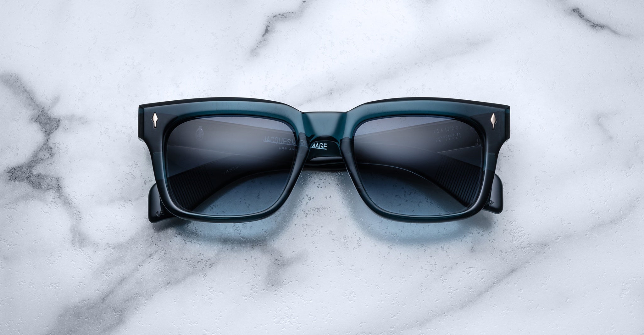 Jacques Marie Mage Torino sunglasses, featuring bold blue square acetate frames and dark lenses, rest on a white marble surface, showcasing the signature craftsmanship of Jacques Marie Mage.