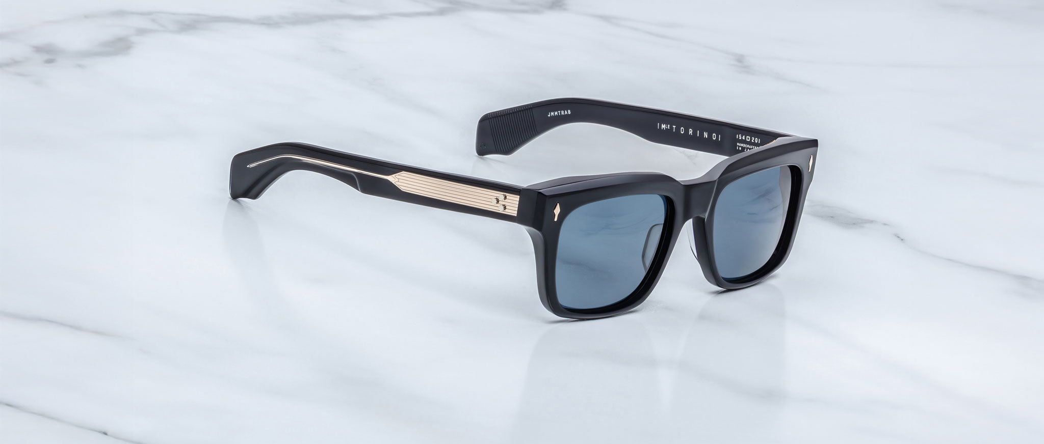 The Jacques Marie Mage Torino by Jacques Marie Mage features black handcrafted acetate rectangular frames with blue-tinted lenses and gold arm detailing, displayed on a white marble surface.