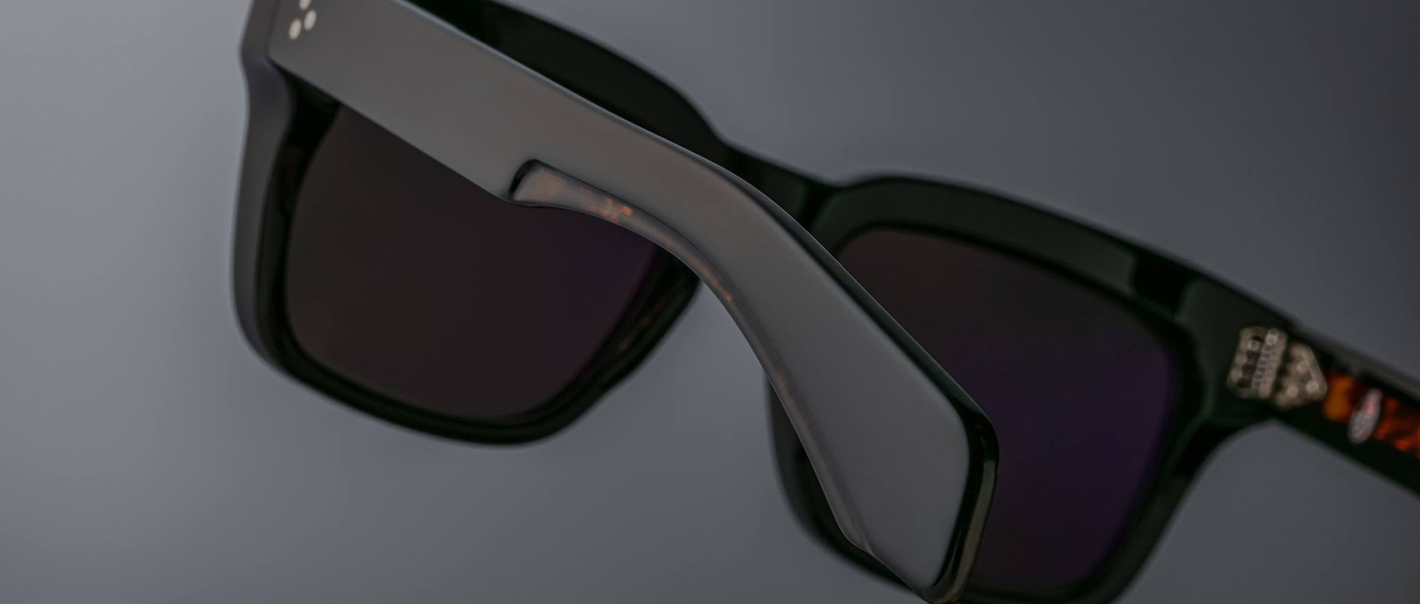 Close-up of Jacques Marie Mage Torino bold rectangular eyeglass frames in black acetate with dark lenses, shown at an angle against a plain gray background.