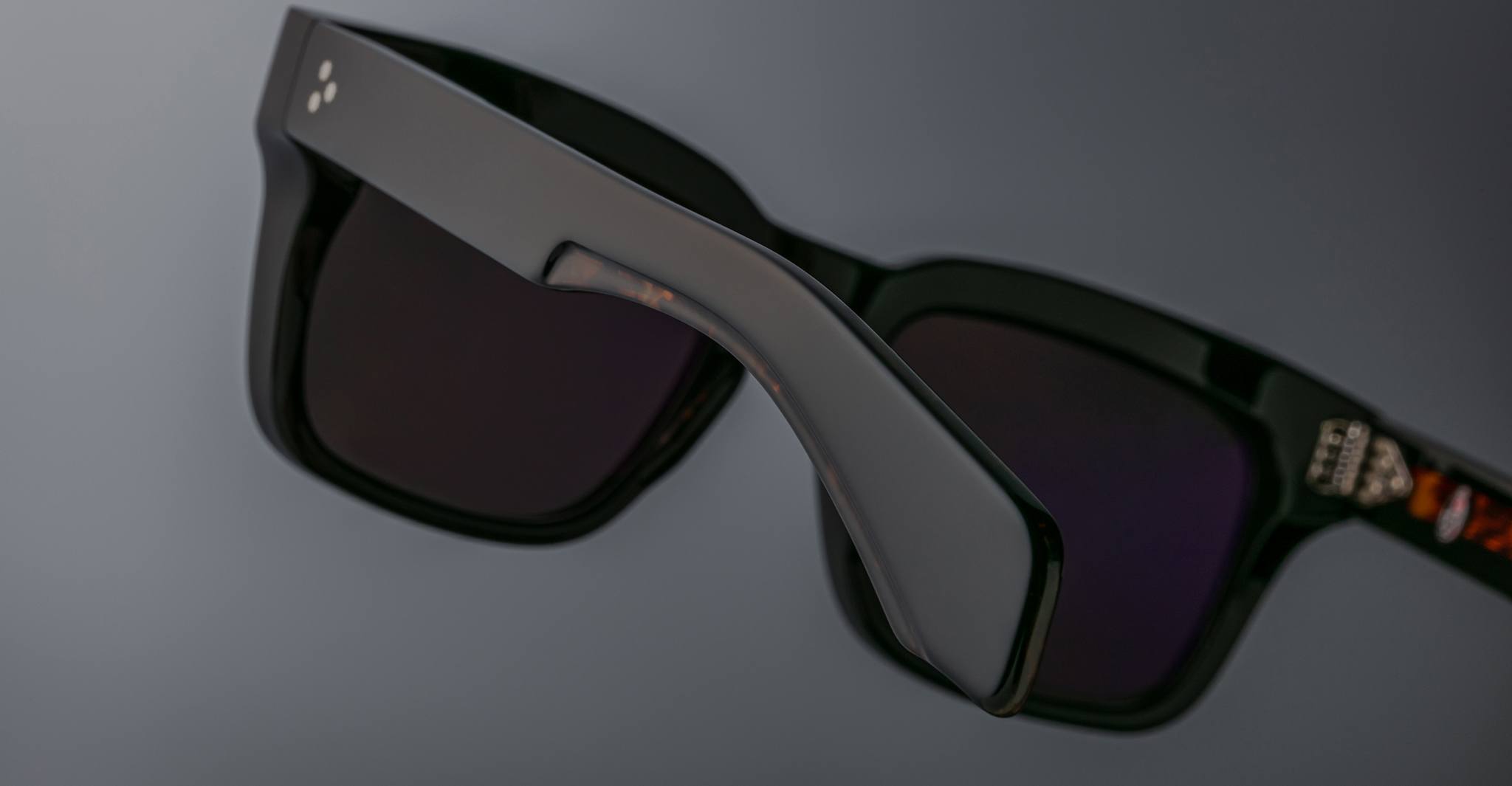 Close-up of Jacques Marie Mage Torino bold rectangular eyeglass frames in black acetate with dark lenses, shown at an angle against a plain gray background.