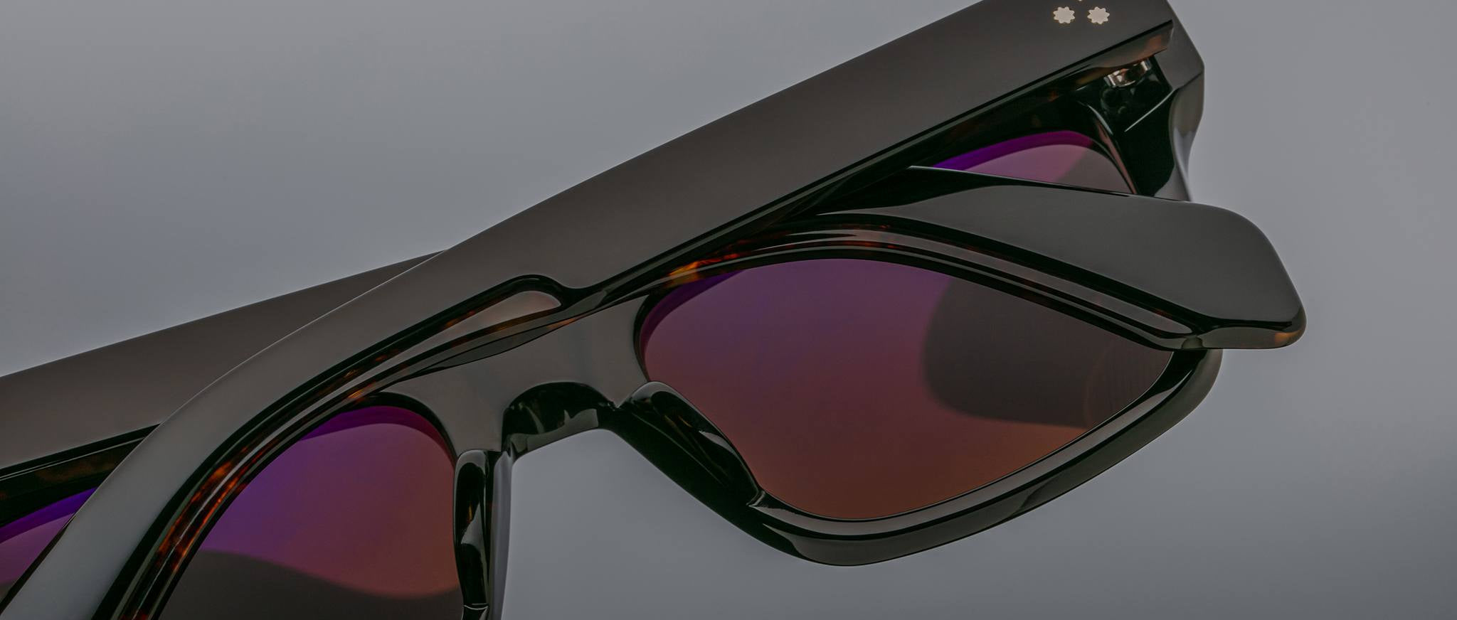 Close-up of Jacques Marie Mage Torino rectangular eyeglass frames, featuring dark tinted lenses and a subtle decorative accent on handcrafted acetate, inspired by the iconic style of Jacques Marie Mage, set against a gray background.