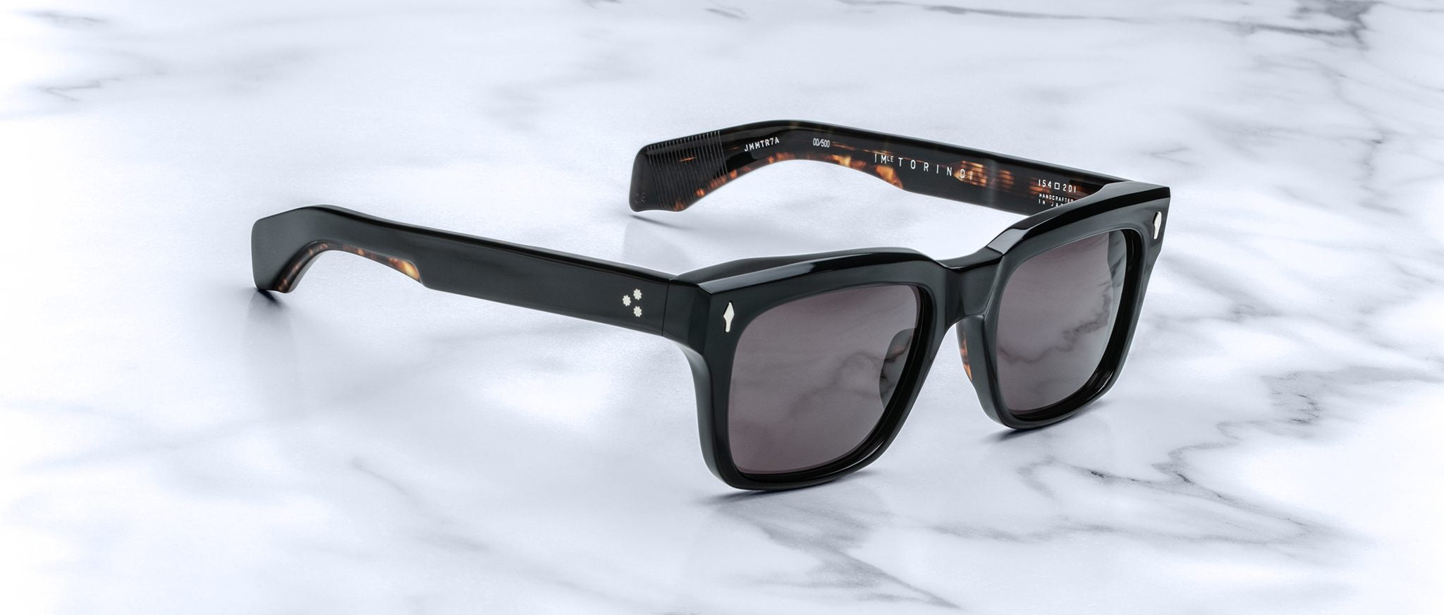 The Jacques Marie Mage Torino sunglasses feature bold black rectangular acetate frames and dark lenses, displayed on a white marble surface to highlight the sophisticated, handcrafted design of Jacques Marie Mage eyewear.