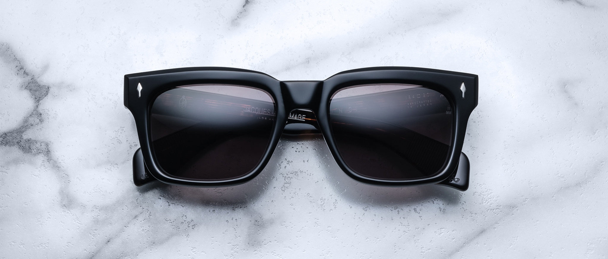 Jacques Marie Mage Torino sunglasses by Jacques Marie Mage feature bold black rectangular acetate frames and dark lenses, displayed on a white marble surface, recalling the handcrafted elegance of the Torino style.