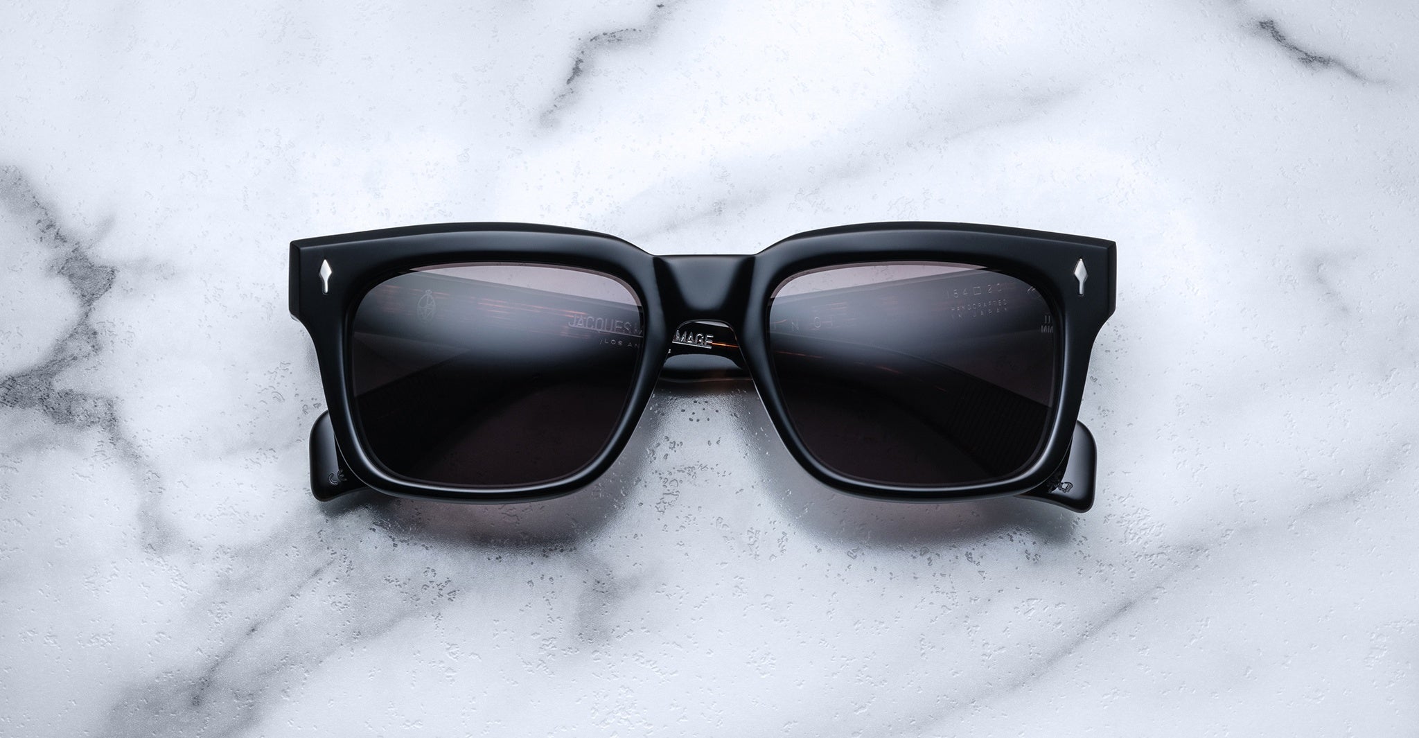 Jacques Marie Mage Torino sunglasses by Jacques Marie Mage feature bold black rectangular acetate frames and dark lenses, displayed on a white marble surface, recalling the handcrafted elegance of the Torino style.