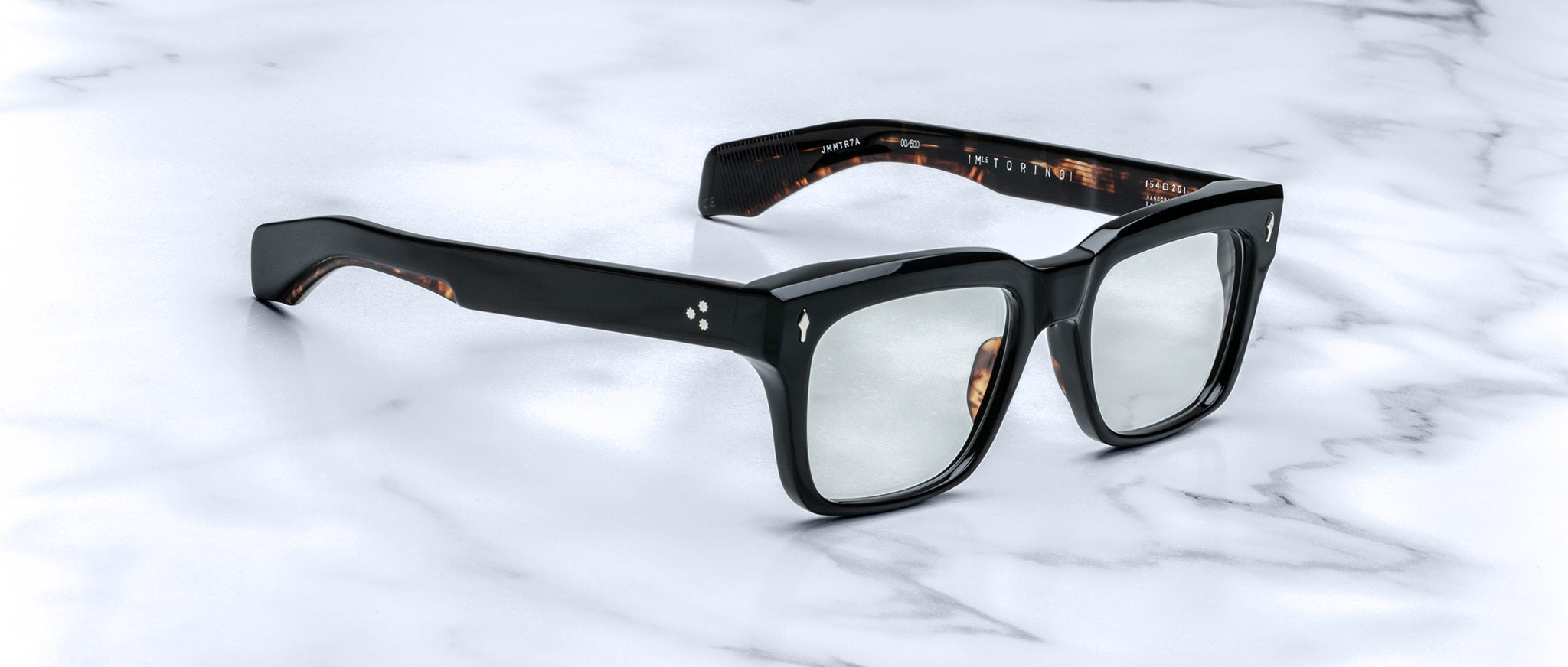 A pair of Jacques Marie Mage Torino black rectangular eyeglass frames by Jacques Marie Mage rests on a white marble surface.