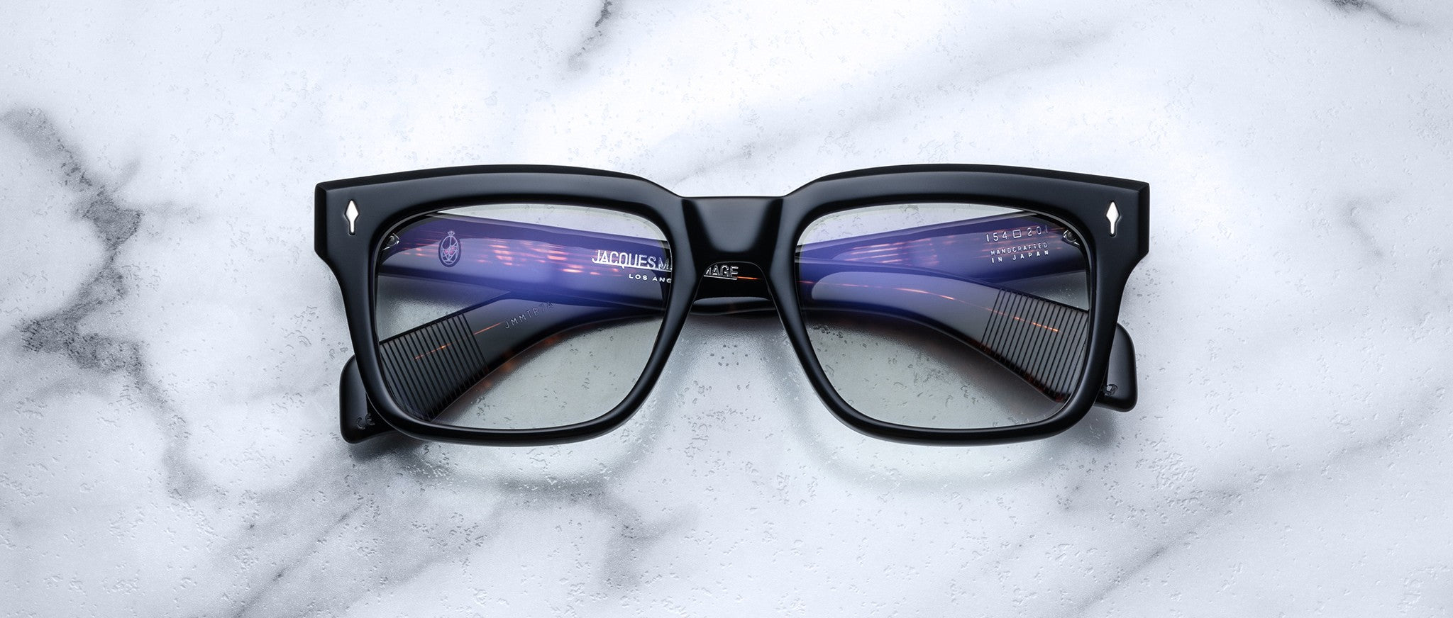 Jacques Marie Mage Torino eyeglasses feature bold black rectangular acetate frames with blue-tinted lenses, shown on a white marble surface. The digital screen reflection highlights the handcrafted design by Jacques Marie Mage.