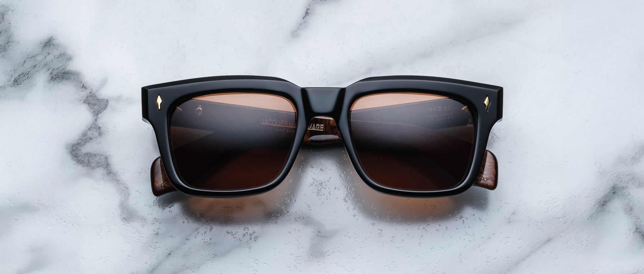Jacques Marie Mage Torino sunglasses feature bold black square acetate frames with brown-tinted lenses, displayed on white marble—a striking example of handcrafted eyewear from Jacques Marie Mage.