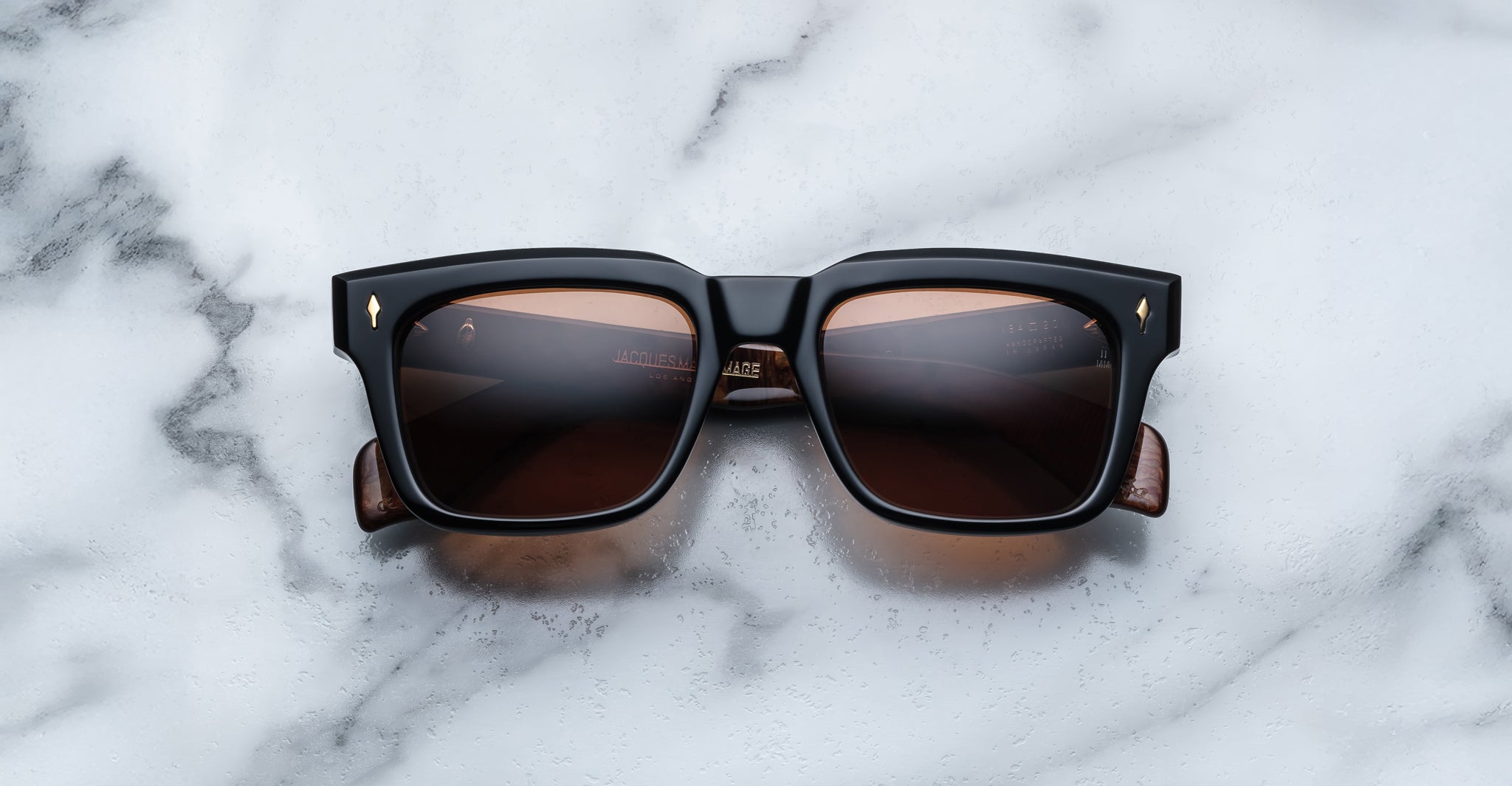 Jacques Marie Mage Torino sunglasses feature bold black square acetate frames with brown-tinted lenses, displayed on white marble—a striking example of handcrafted eyewear from Jacques Marie Mage.