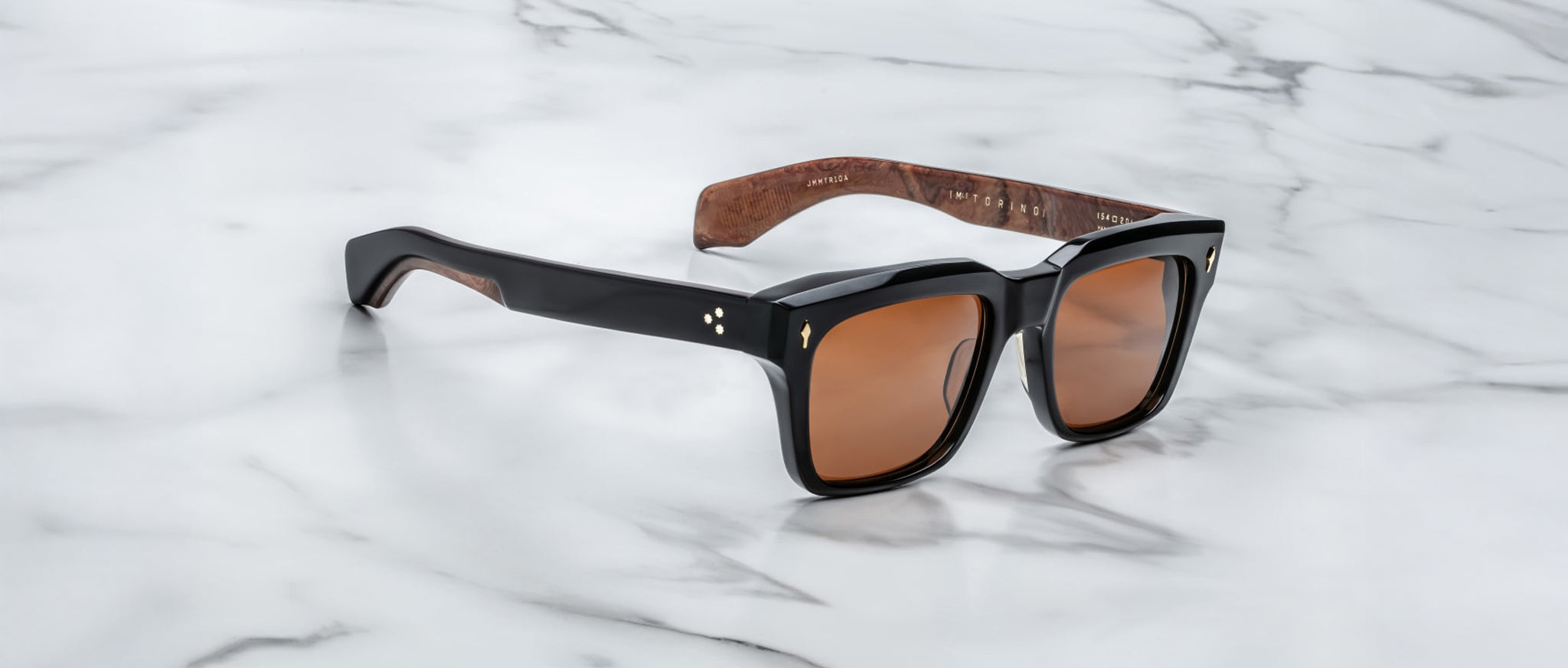 The Jacques Marie Mage Torino features bold black rectangular acetate frames, brown tinted lenses, and wood-textured inner arms, showcased on a white marble surface.
