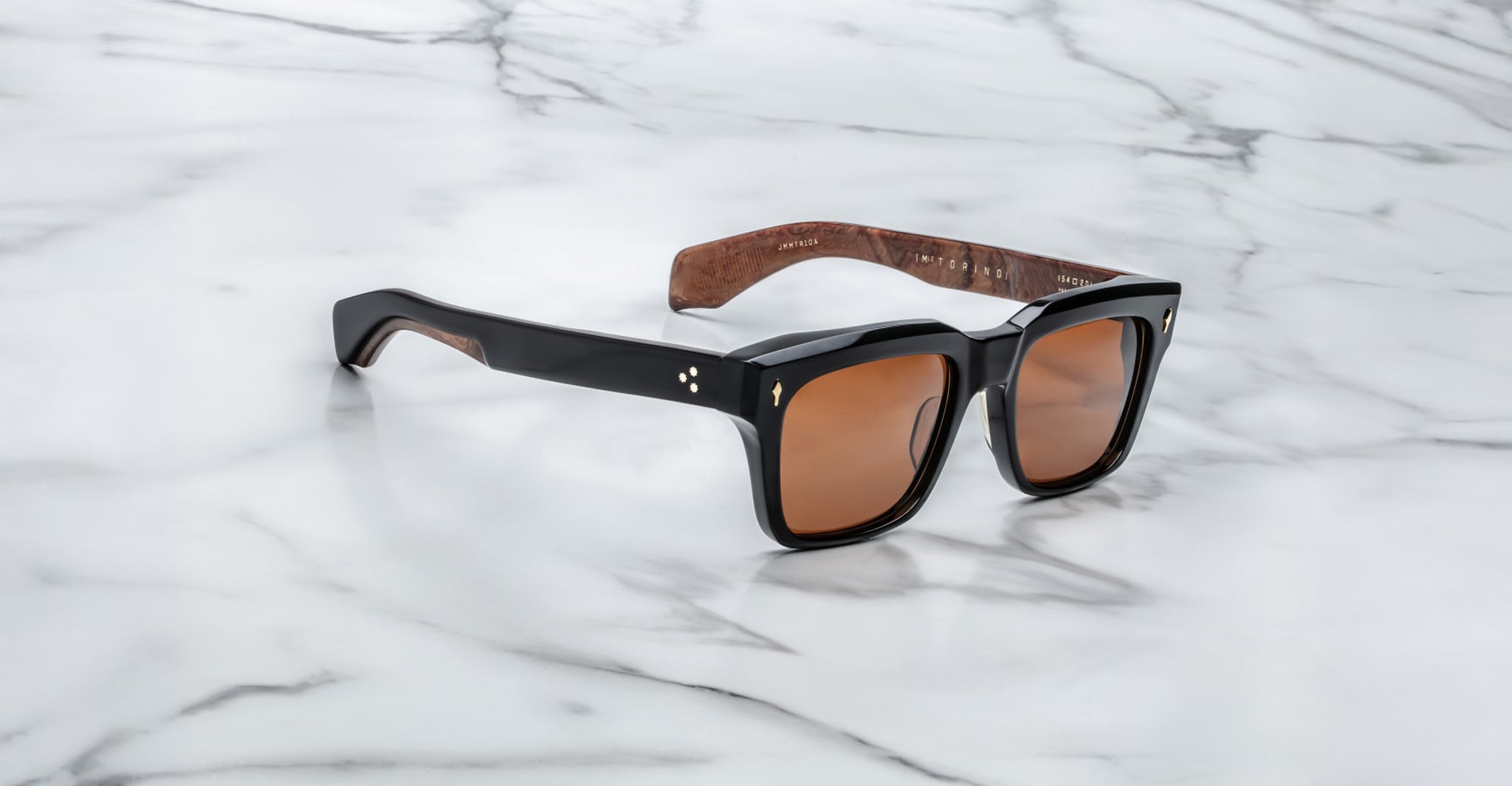 The Jacques Marie Mage Torino features bold black rectangular acetate frames, brown tinted lenses, and wood-textured inner arms, showcased on a white marble surface.